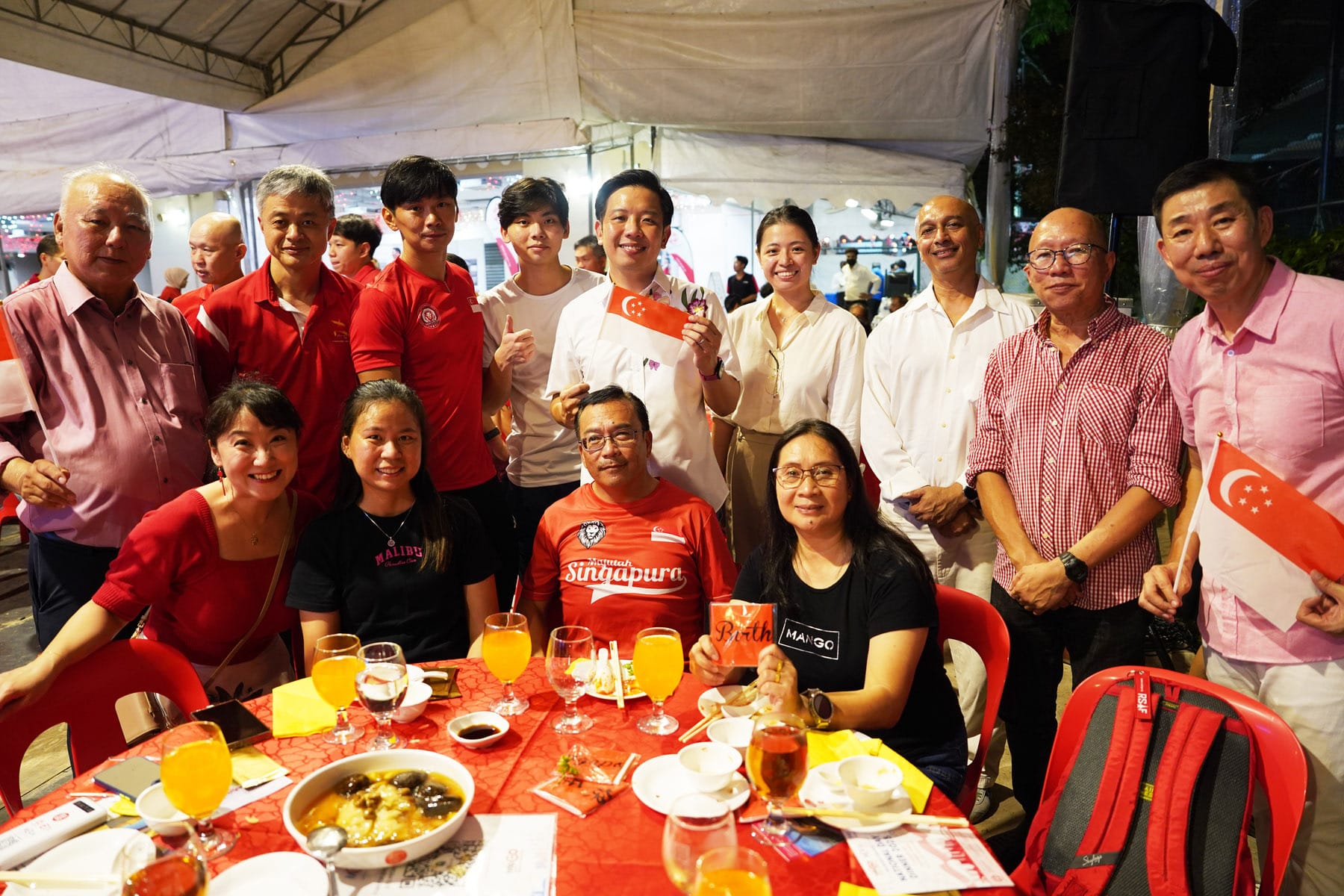 Group of people posing at a dinner table under a tent, holding small Singapore flags, wearing red and white attire.