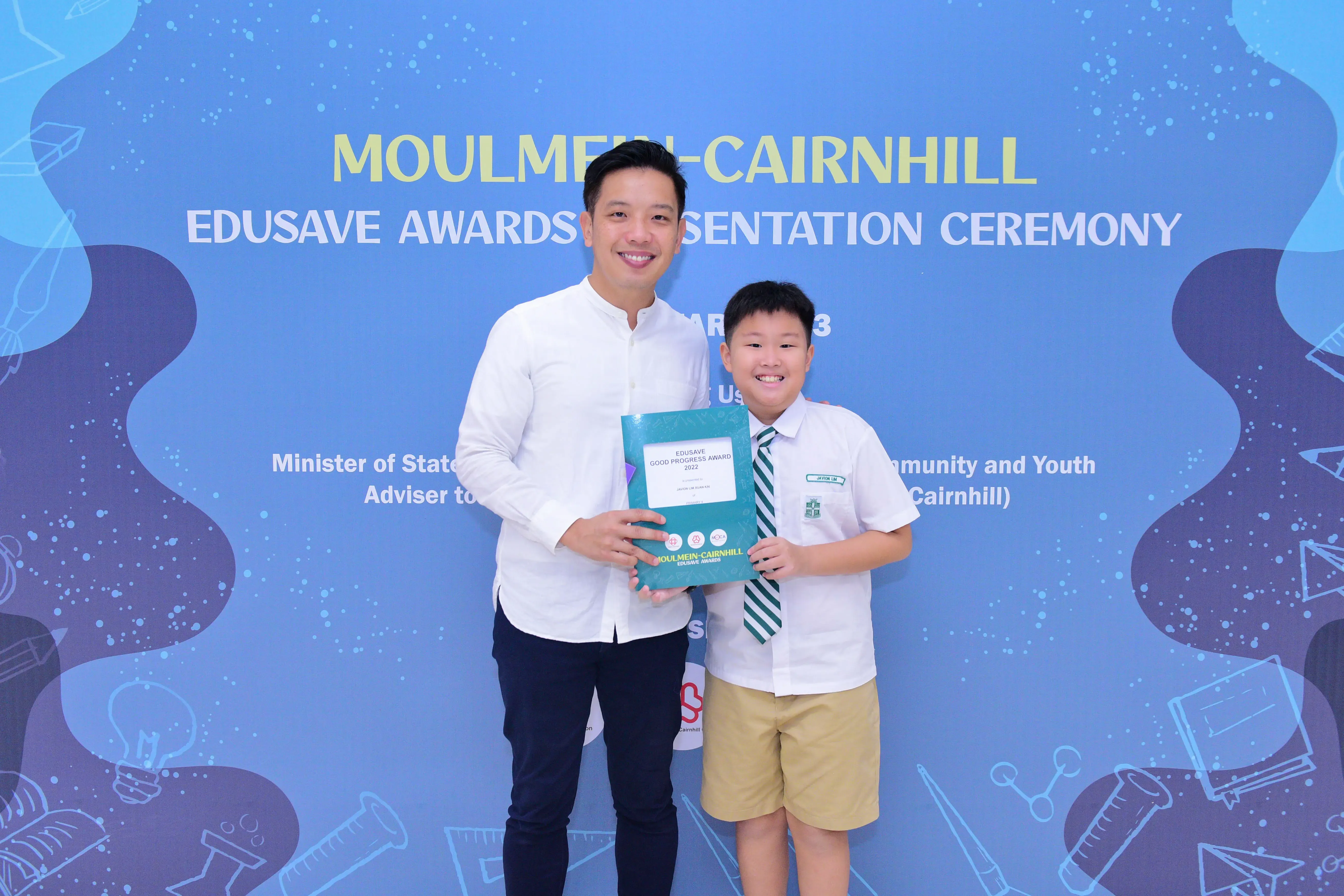 A man and a young boy in school uniform hold an Edusave Good Progress Award at the Moulmein-Cairnhill ceremony.