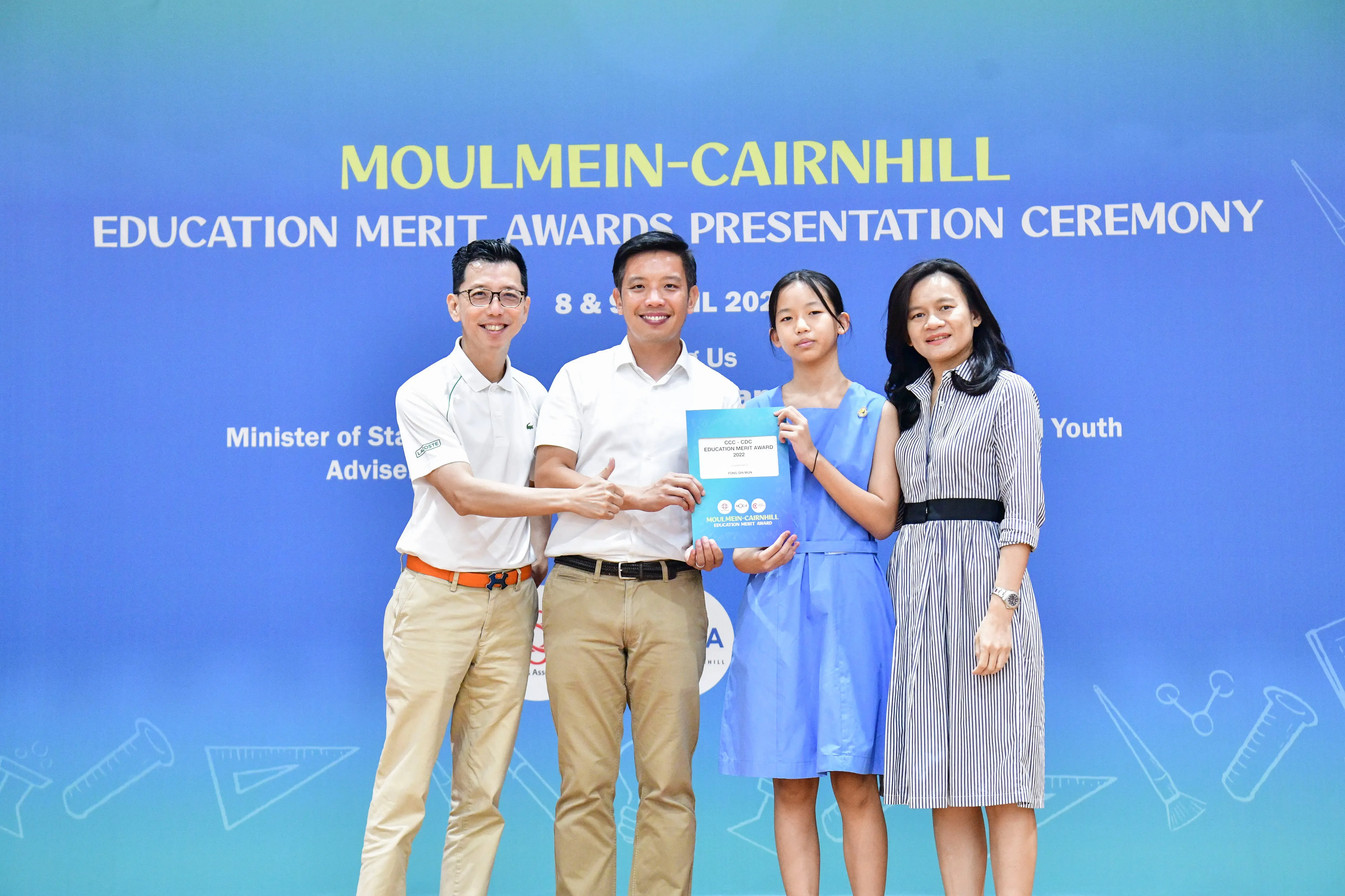 Four people pose with a certificate at the Moulmein-Cairnhill Education Merit Awards Presentation Ceremony banner.