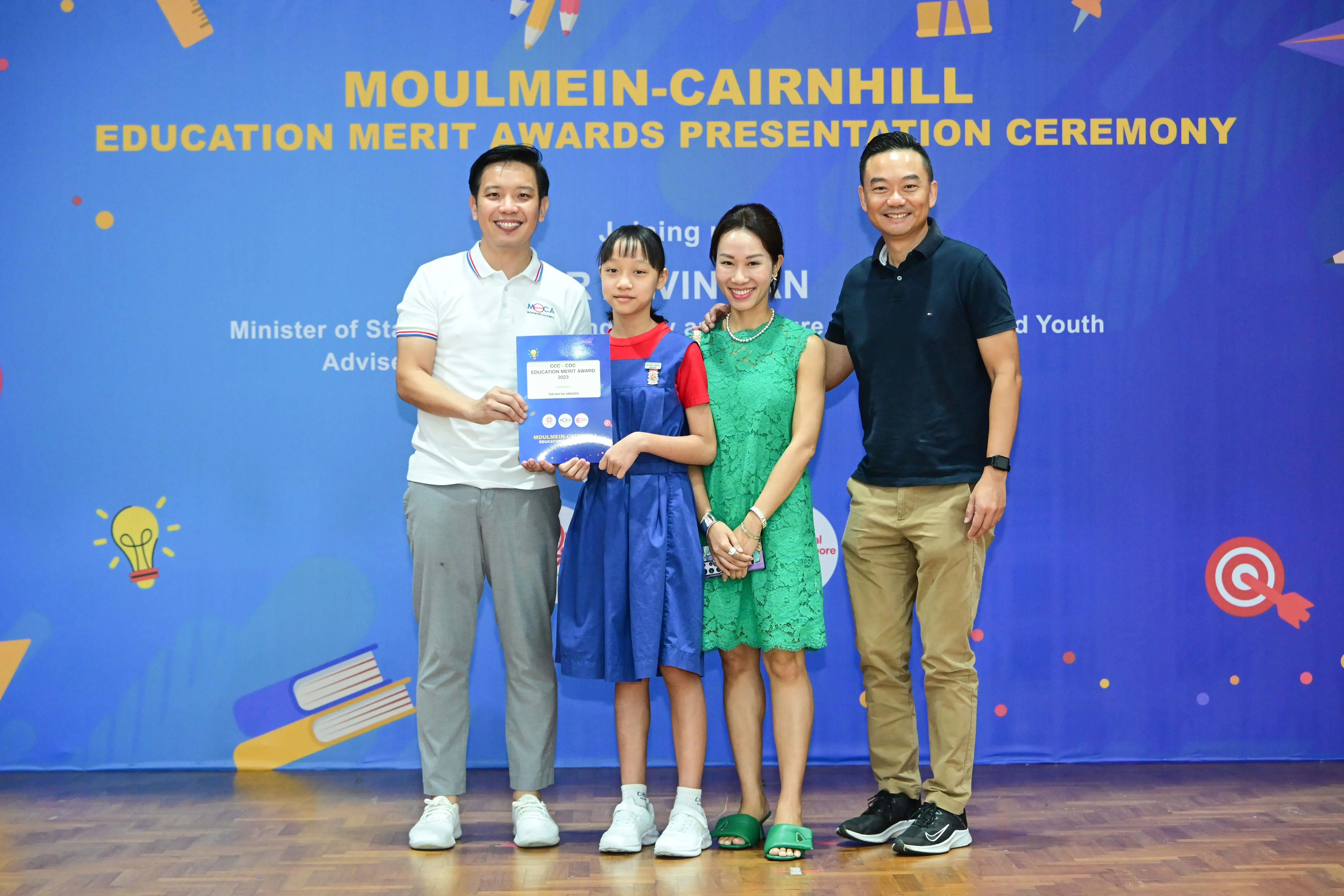 Four people pose at the Moulmein-Cairnhill Education Merit Awards Presentation Ceremony, with a girl holding a certificate.
