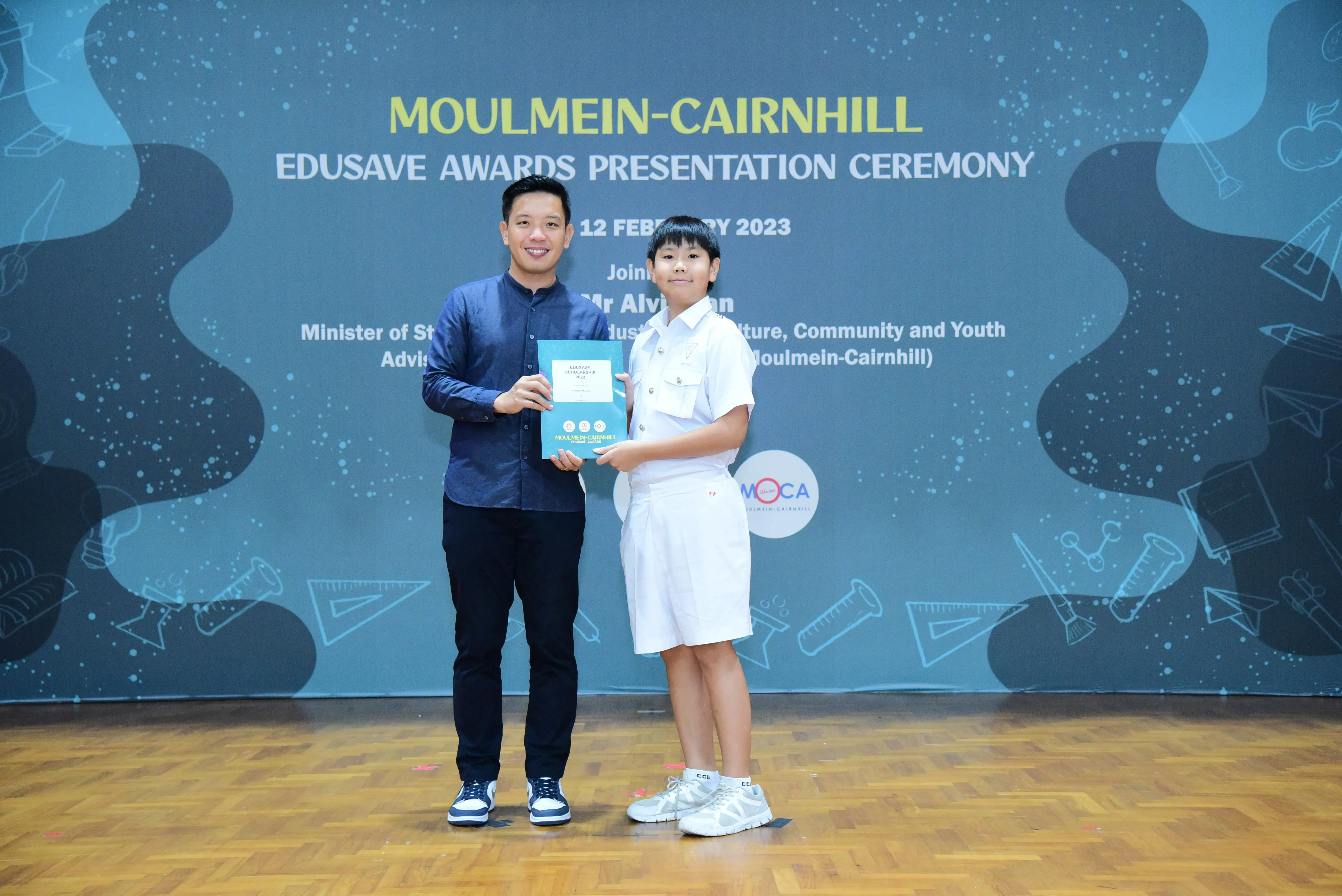 An adult and a young student in white uniform hold a certificate at the Moulmein-Cairnhill Edusave Awards Ceremony 2023.