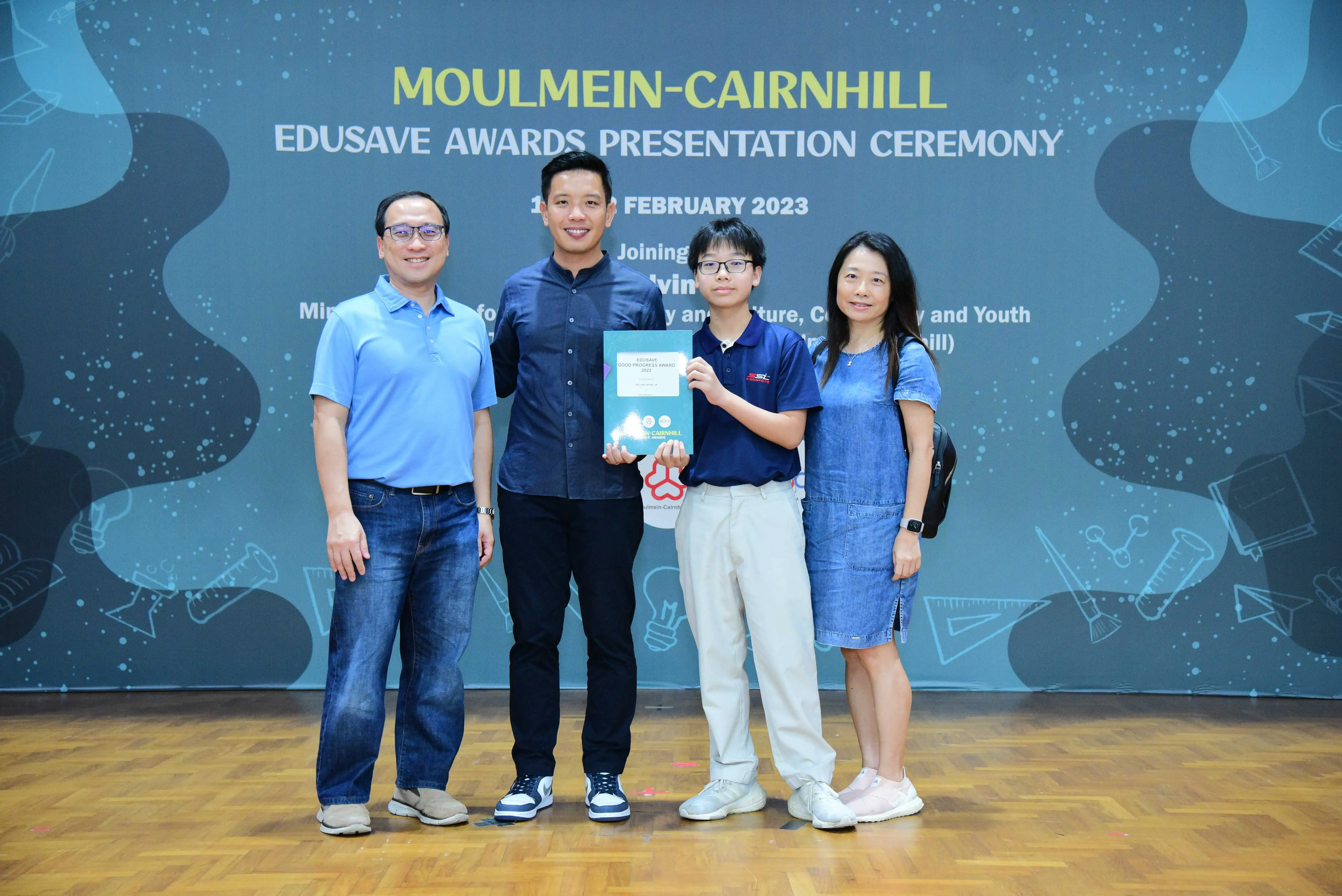 Four people pose in front of a Moulmein-Cairnhill Edusave Awards Presentation Ceremony banner, February 2023.
