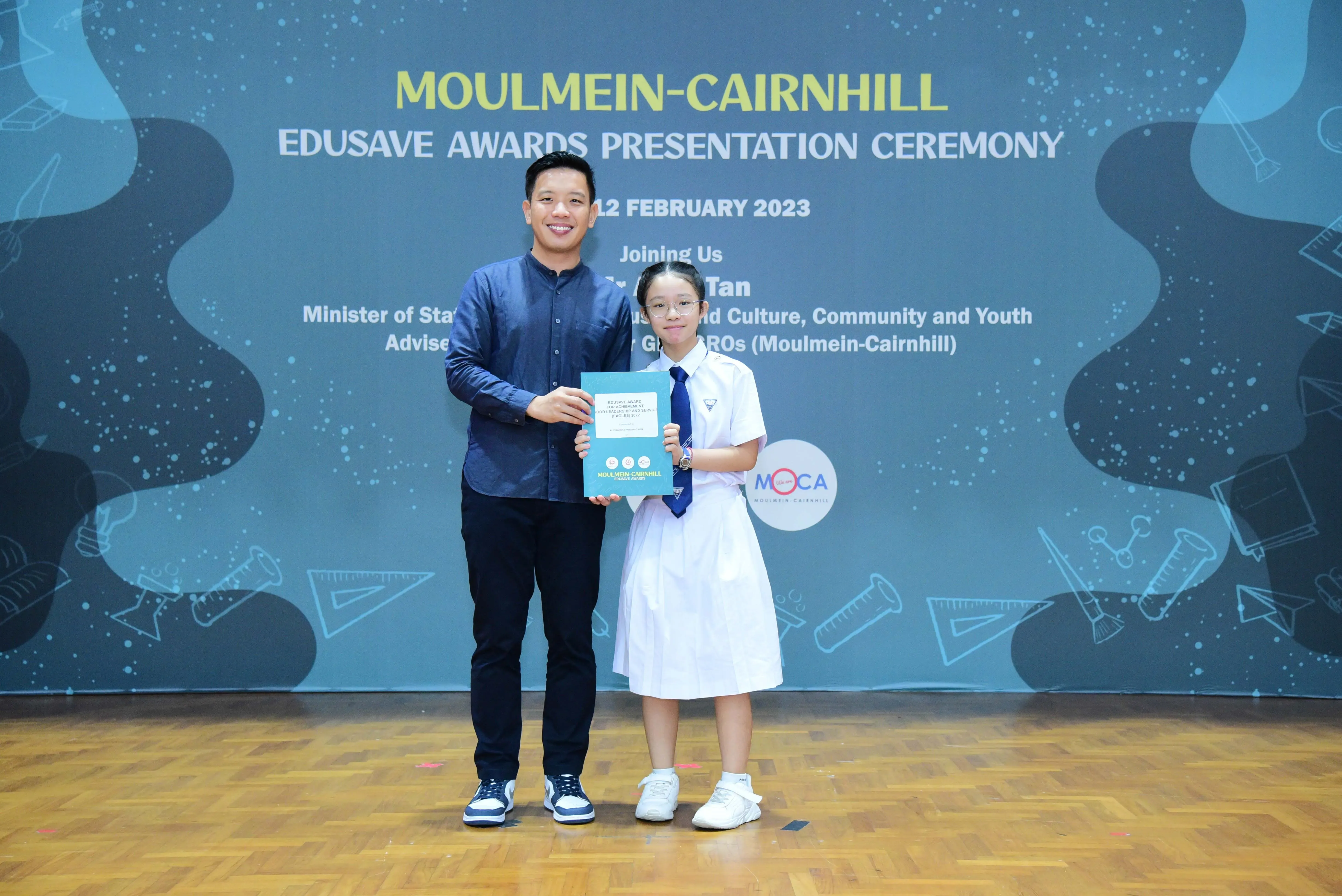 A man and a young girl in school uniform hold a certificate at the Moulmein-Cairnhill Edusave Awards Ceremony, February 2023.