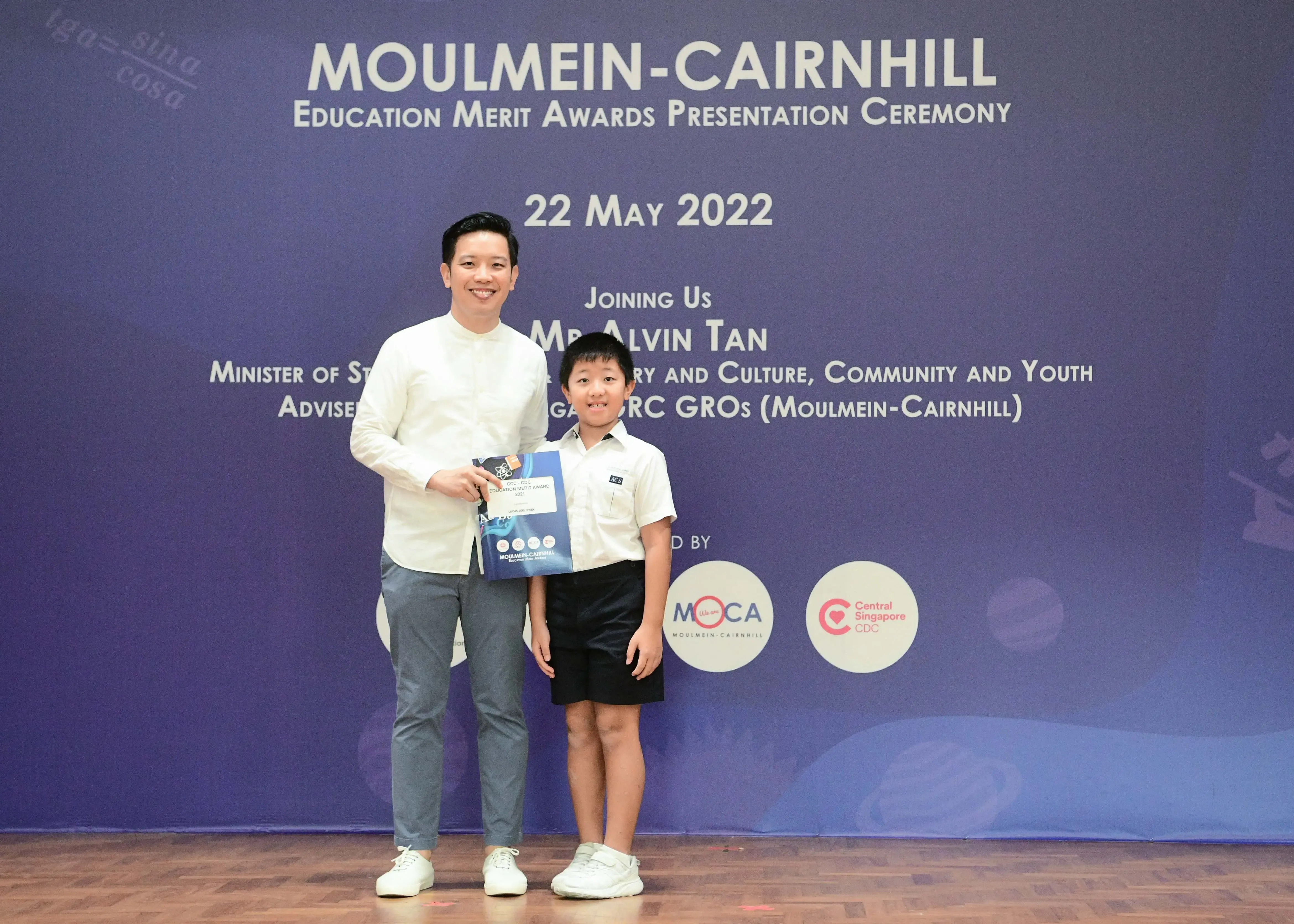 A man and a young boy in school uniform pose with an award at the Moulmein-Cairnhill Education Merit Awards ceremony.