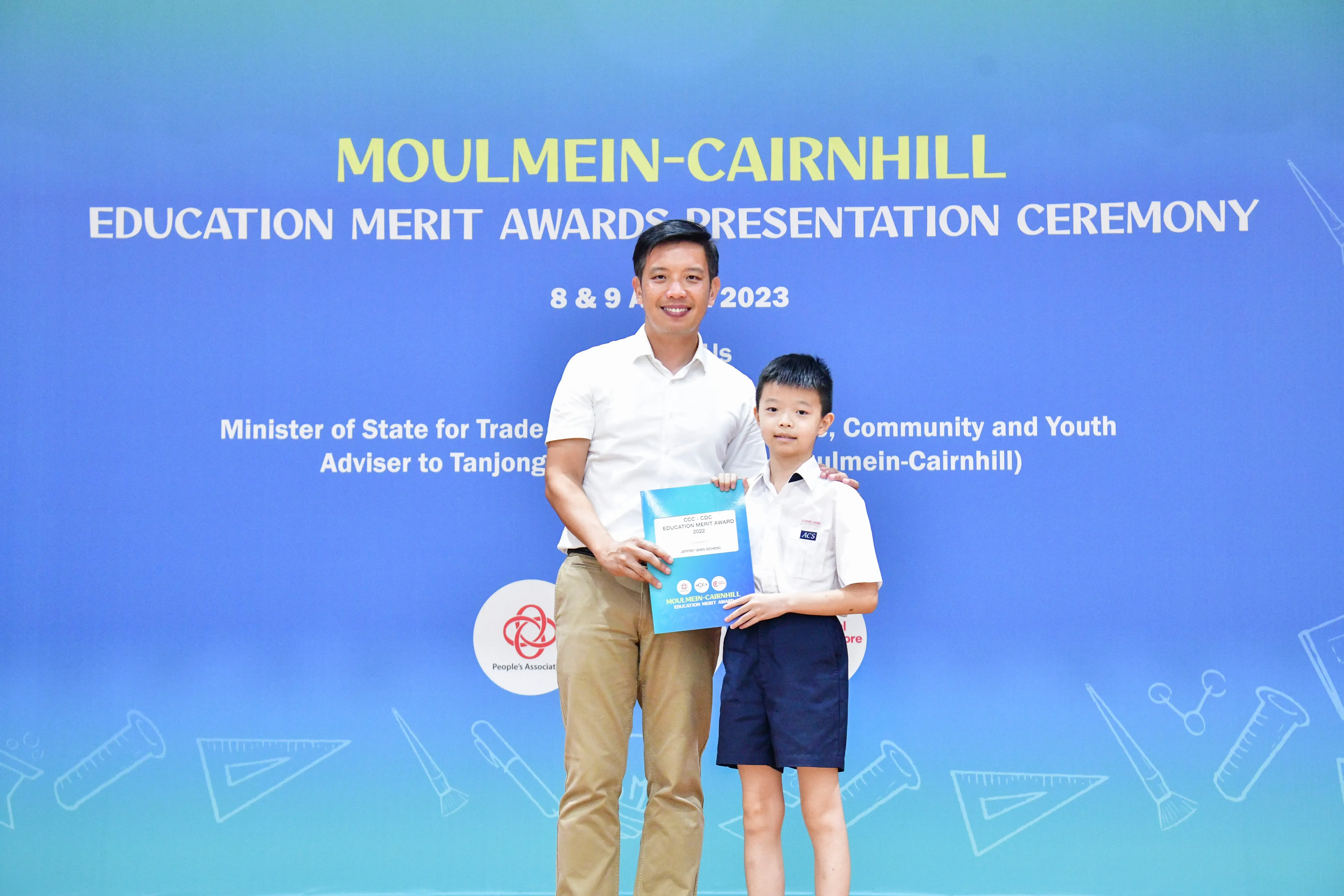 A man and a young boy hold an award certificate in front of a Moulmein-Cairnhill Education Merit Awards banner.