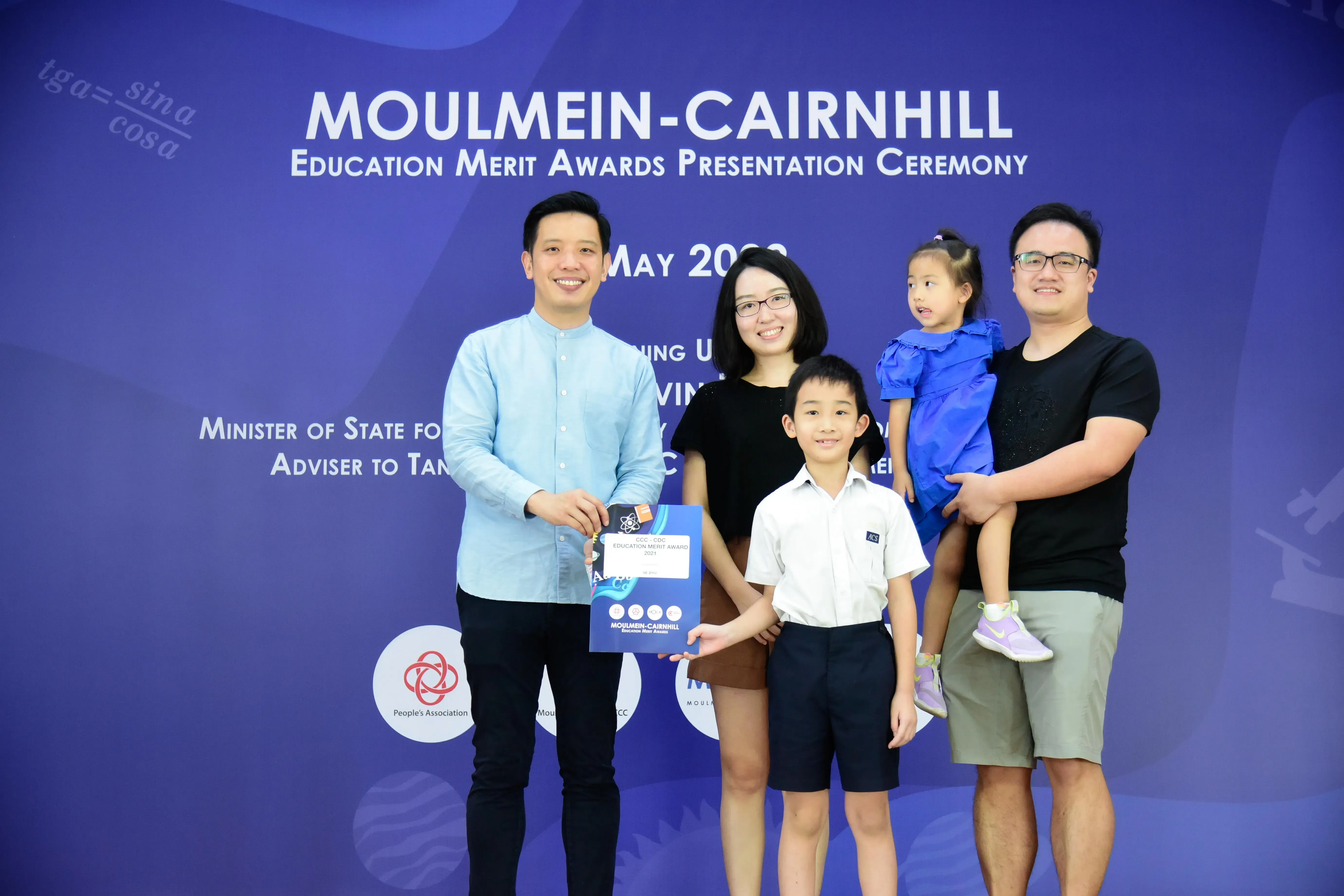 Family of four poses with an official holding a certificate at Moulmein-Cairnhill Education Merit Awards ceremony.