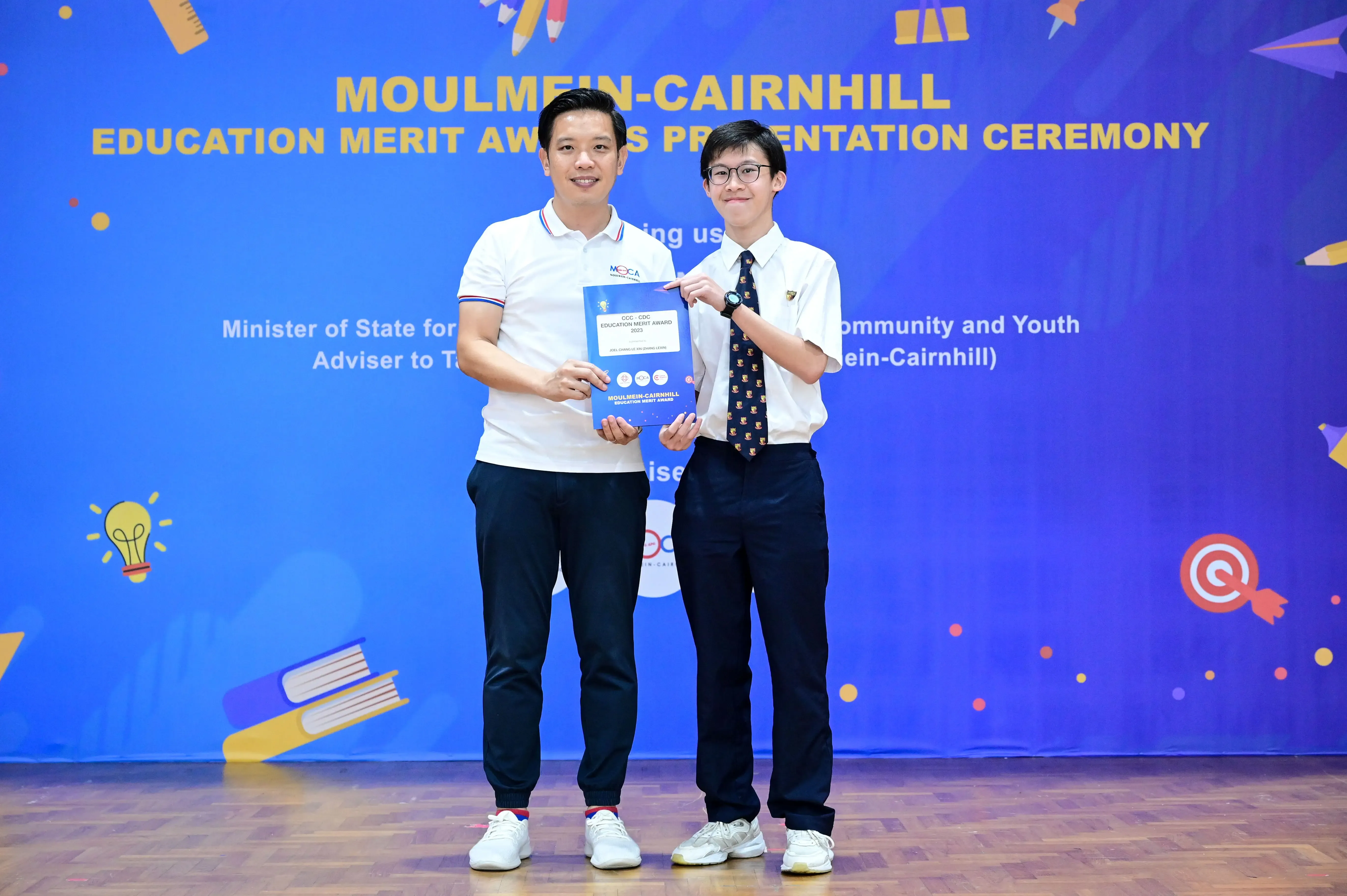 Two people hold a certificate together in front of a blue Moulmein-Cairnhill Education Merit Awards banner.