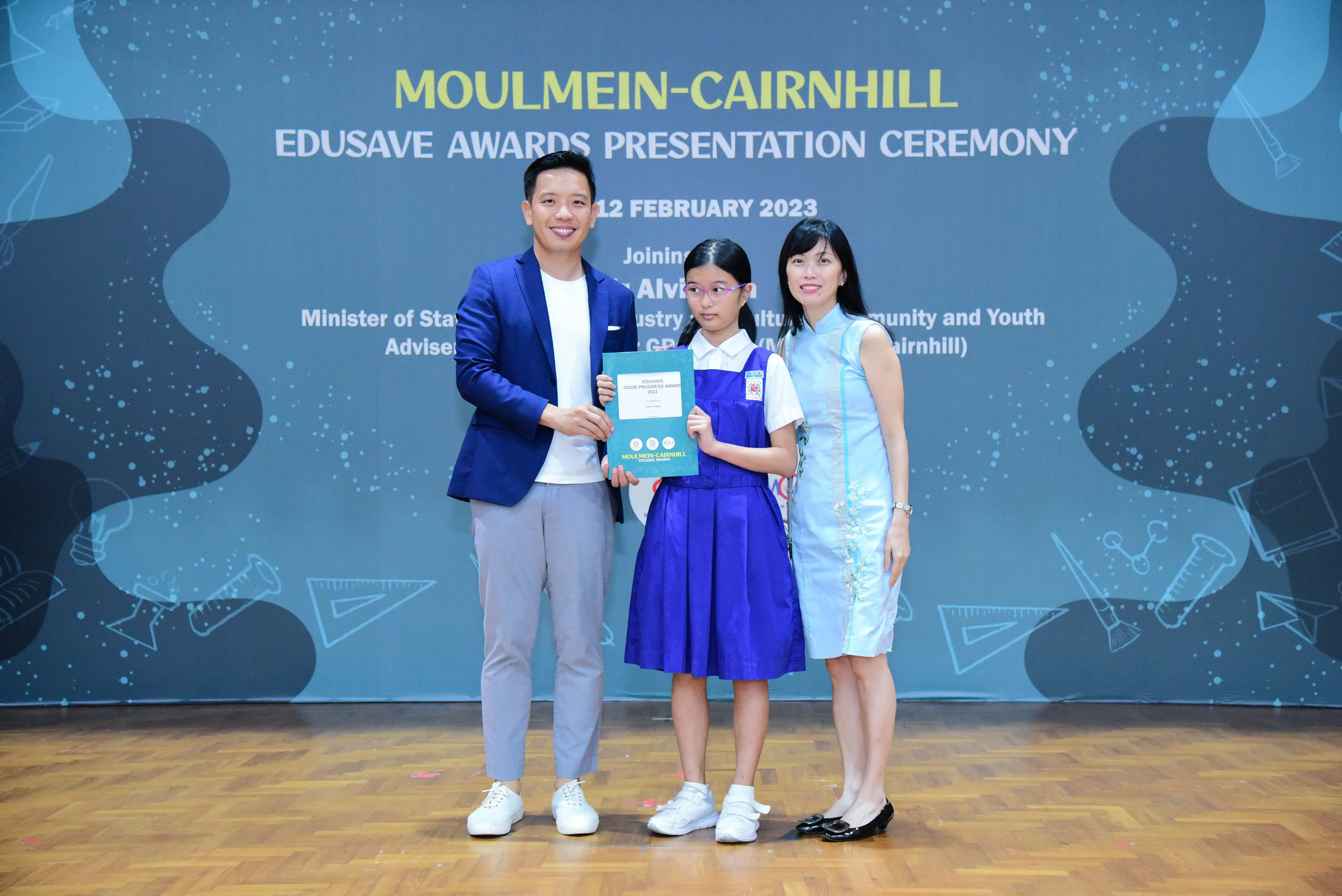 A girl in school uniform holds a certificate between two adults at the Moulmein-Cairnhill Edusave Awards Ceremony, February 2023.