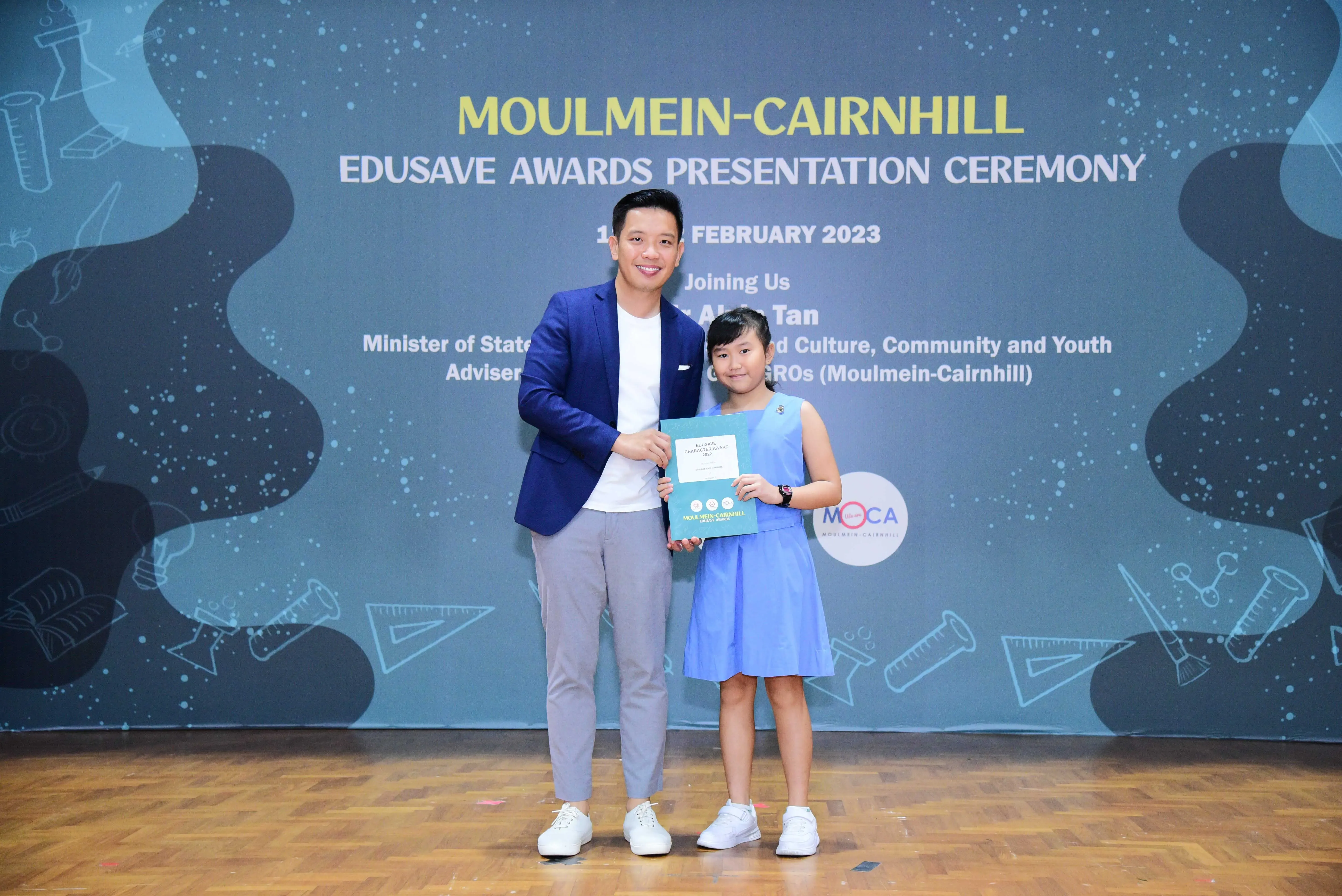 A man in a blue blazer and a young girl in a blue dress hold a certificate at the Moulmein-Cairnhill Edusave Awards Ceremony 2023.