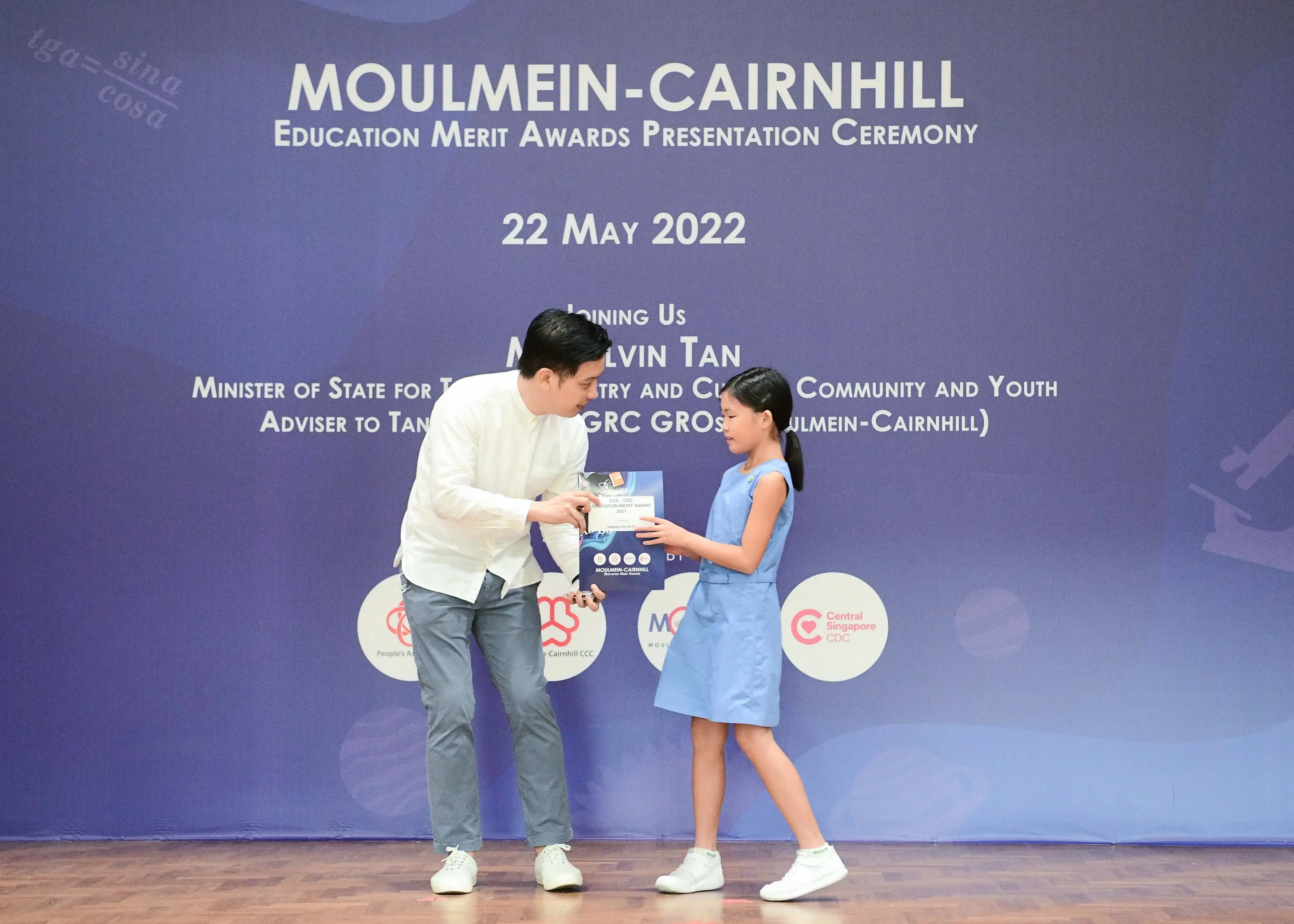 A man in white hands an award to a young girl in a blue dress on stage at the Moulmein-Cairnhill ceremony.