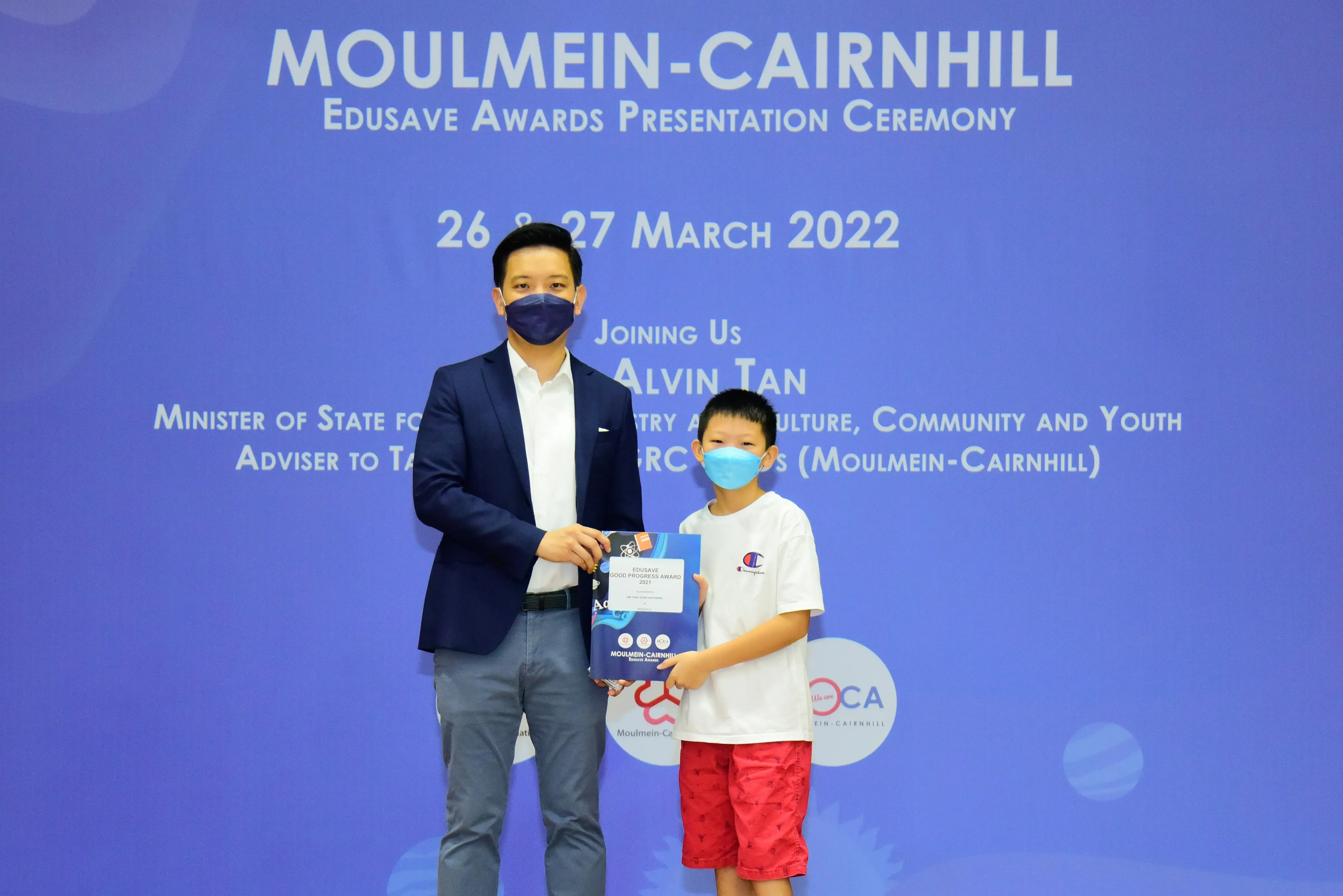 An adult and a young boy wearing masks hold an award at the Moulmein-Cairnhill Edusave Awards Ceremony, March 2022.