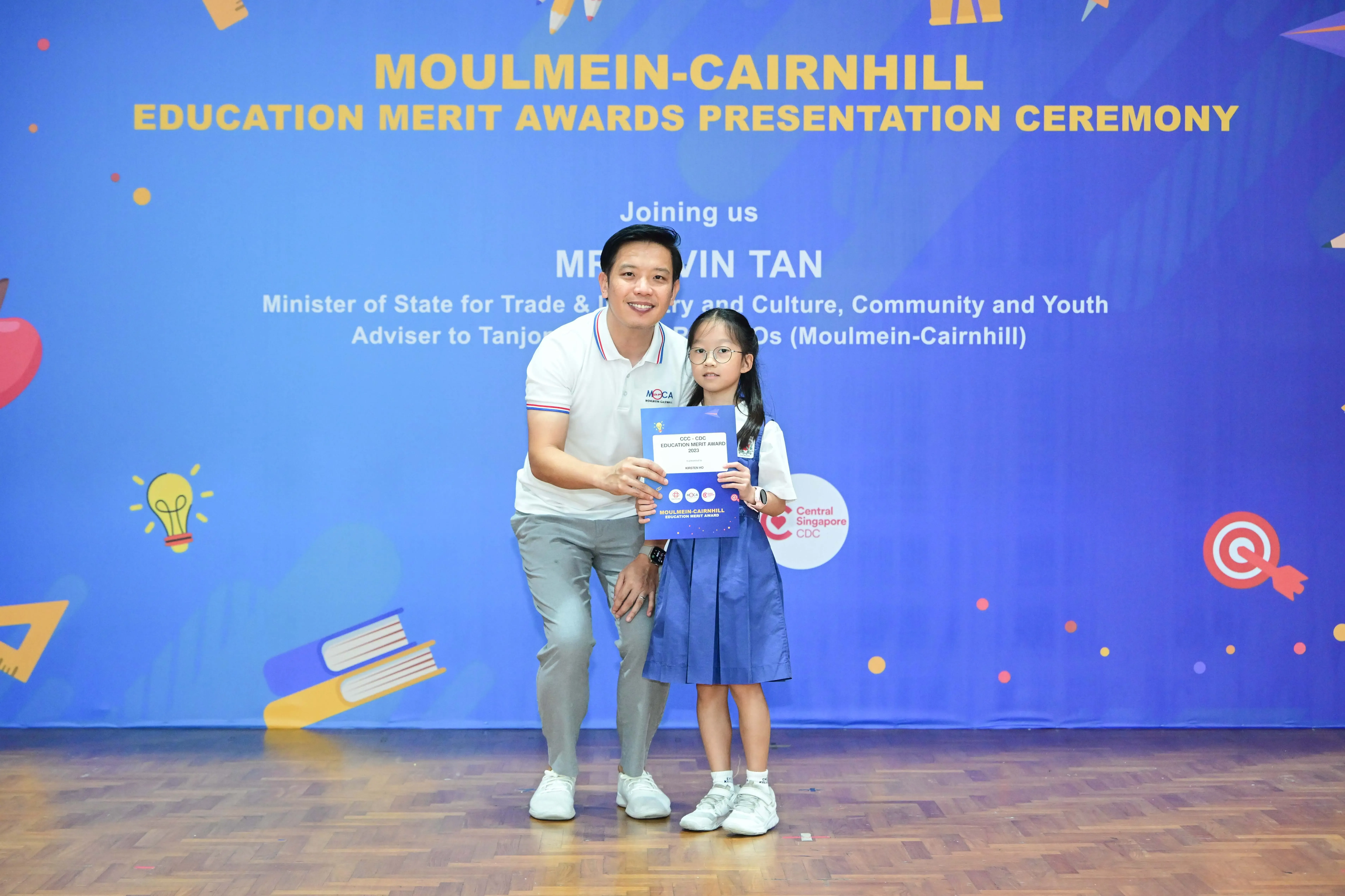 Man and young girl hold an award certificate in front of a Moulmein-Cairnhill Education Merit Awards banner.