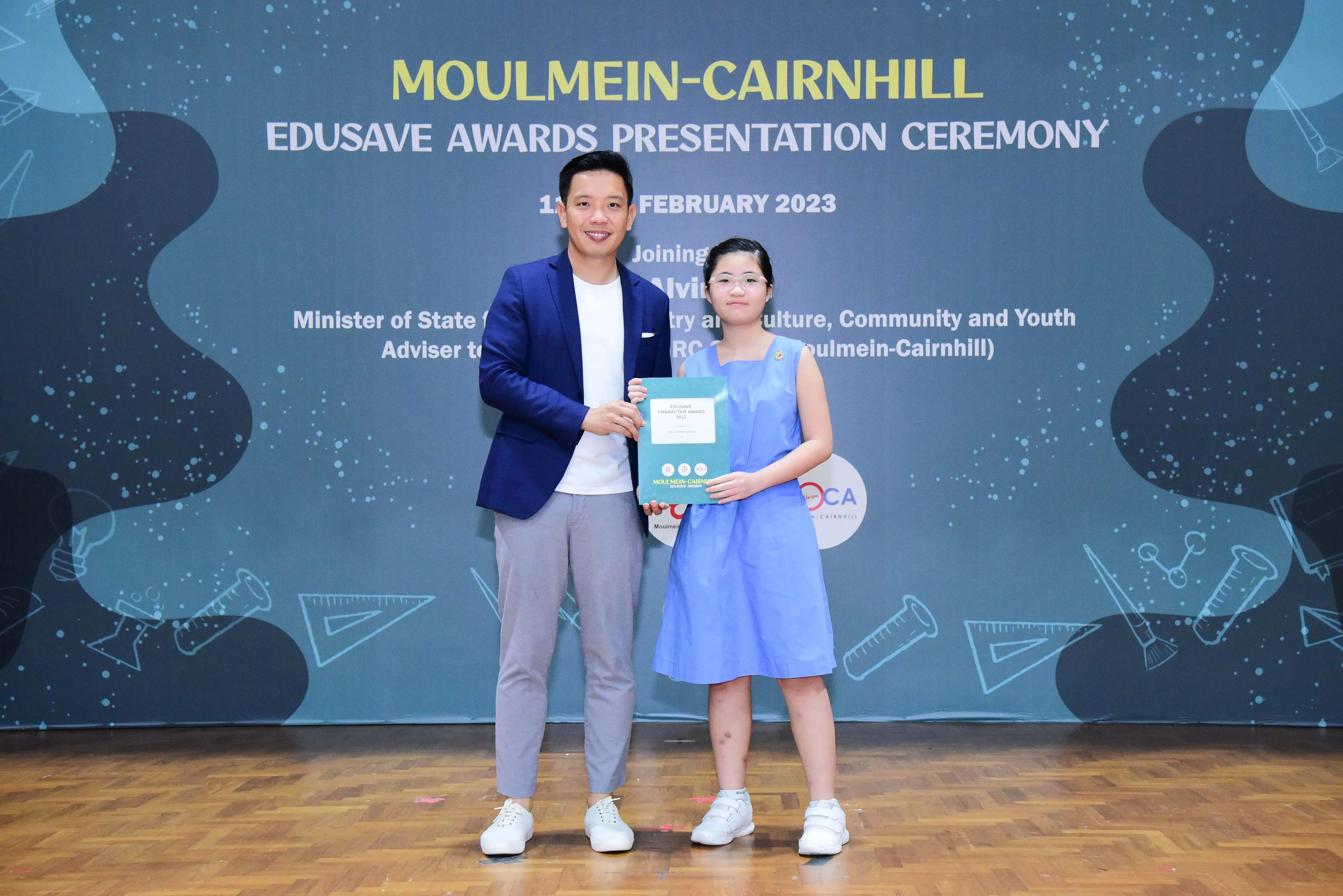 A man in a blue blazer and a young girl in a blue dress hold a certificate at the Moulmein-Cairnhill Edusave Awards Ceremony 2023.