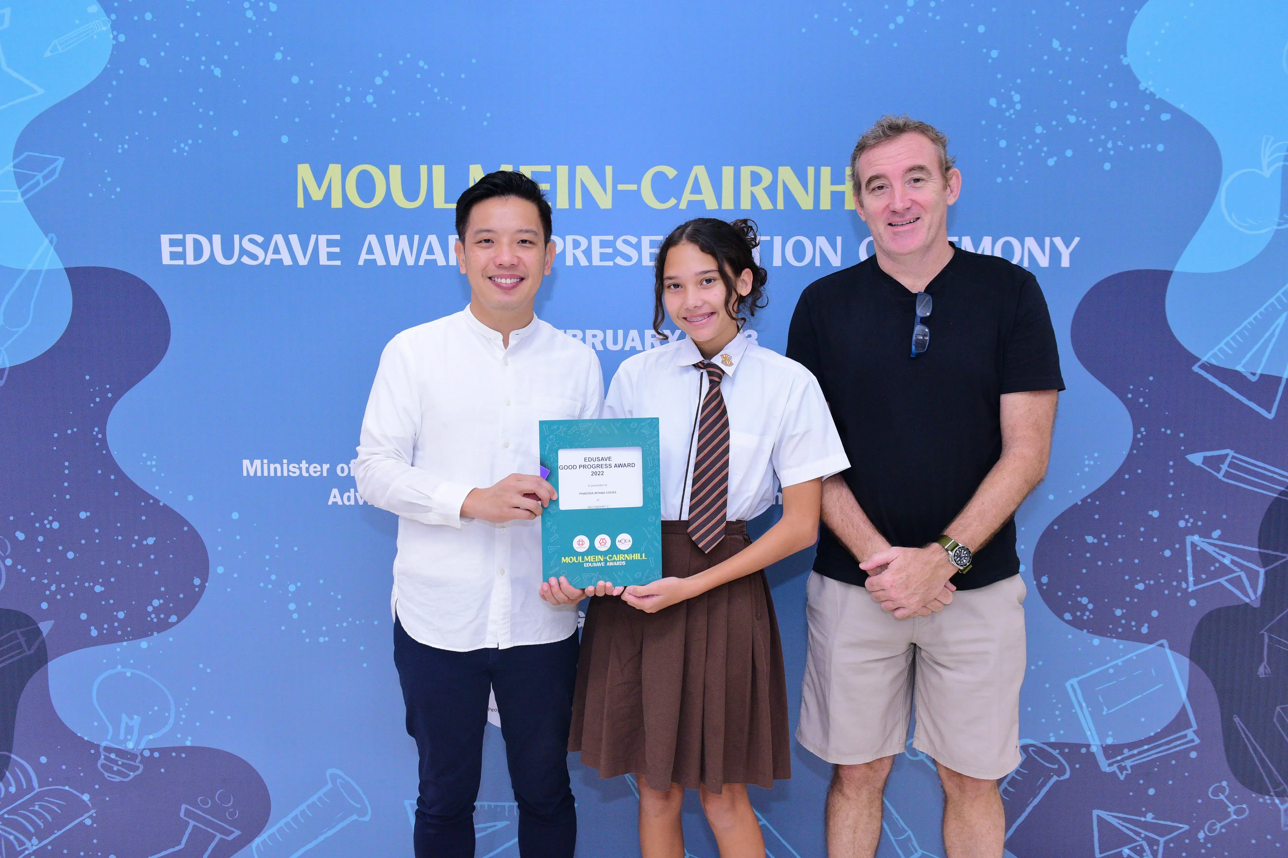 Three people pose at a Moulmein-Cairnhill Edusave Award Presentation Ceremony, with a student holding an award certificate.