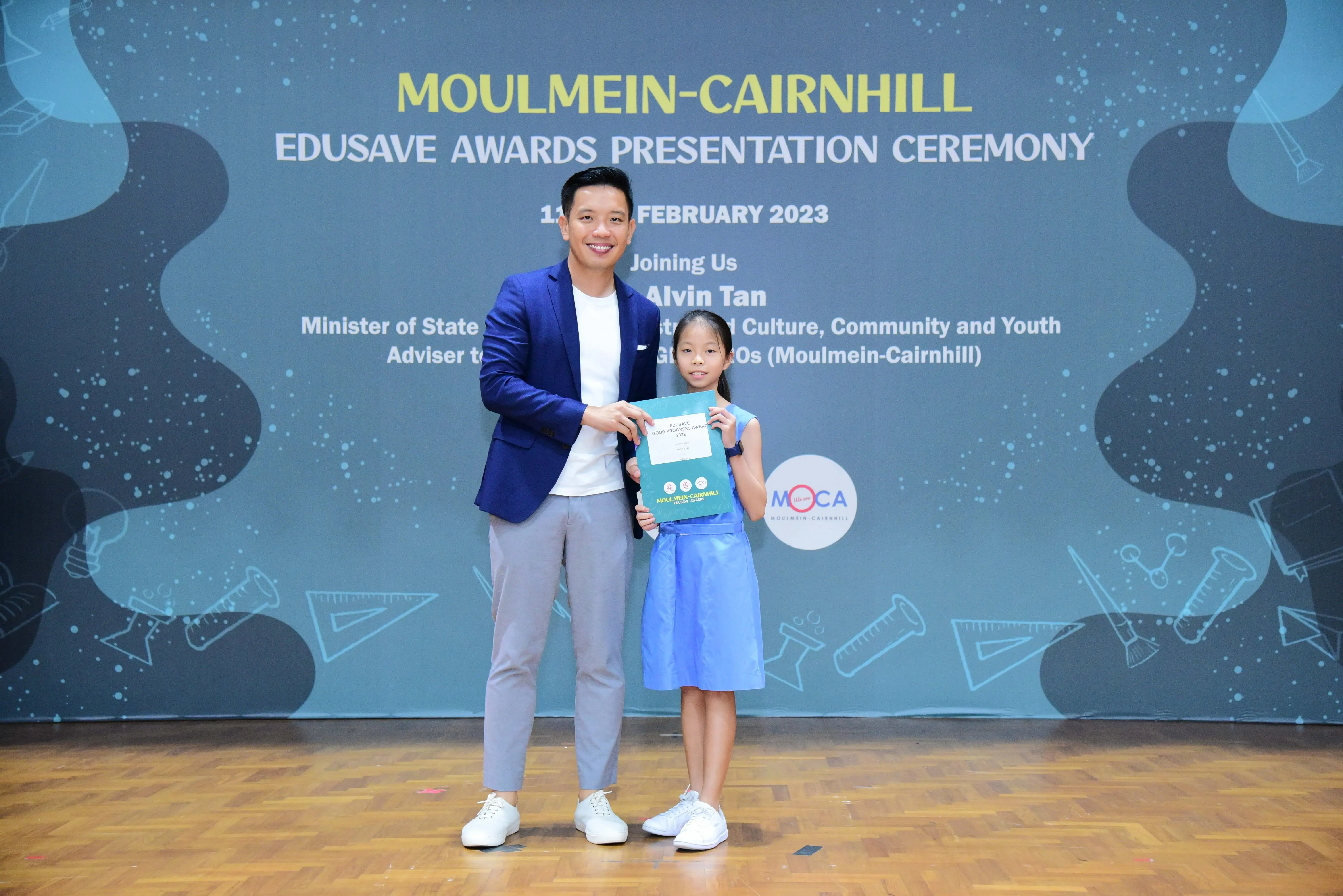 A man in a blue blazer presents a certificate to a young girl at the Moulmein-Cairnhill Edusave Awards Ceremony 2023.