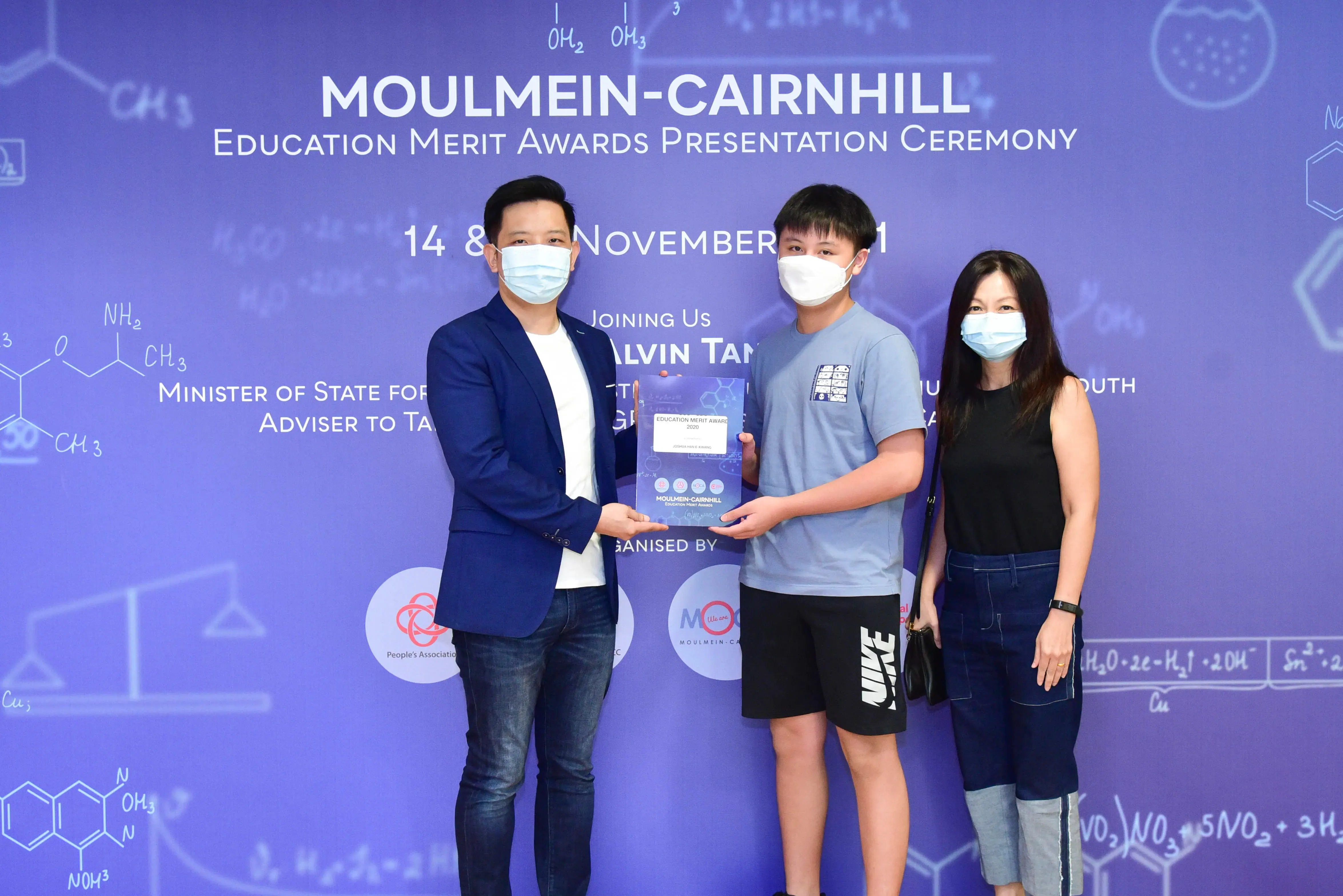 Three masked people pose at the Moulmein-Cairnhill Education Merit Awards Presentation Ceremony, holding an award plaque.