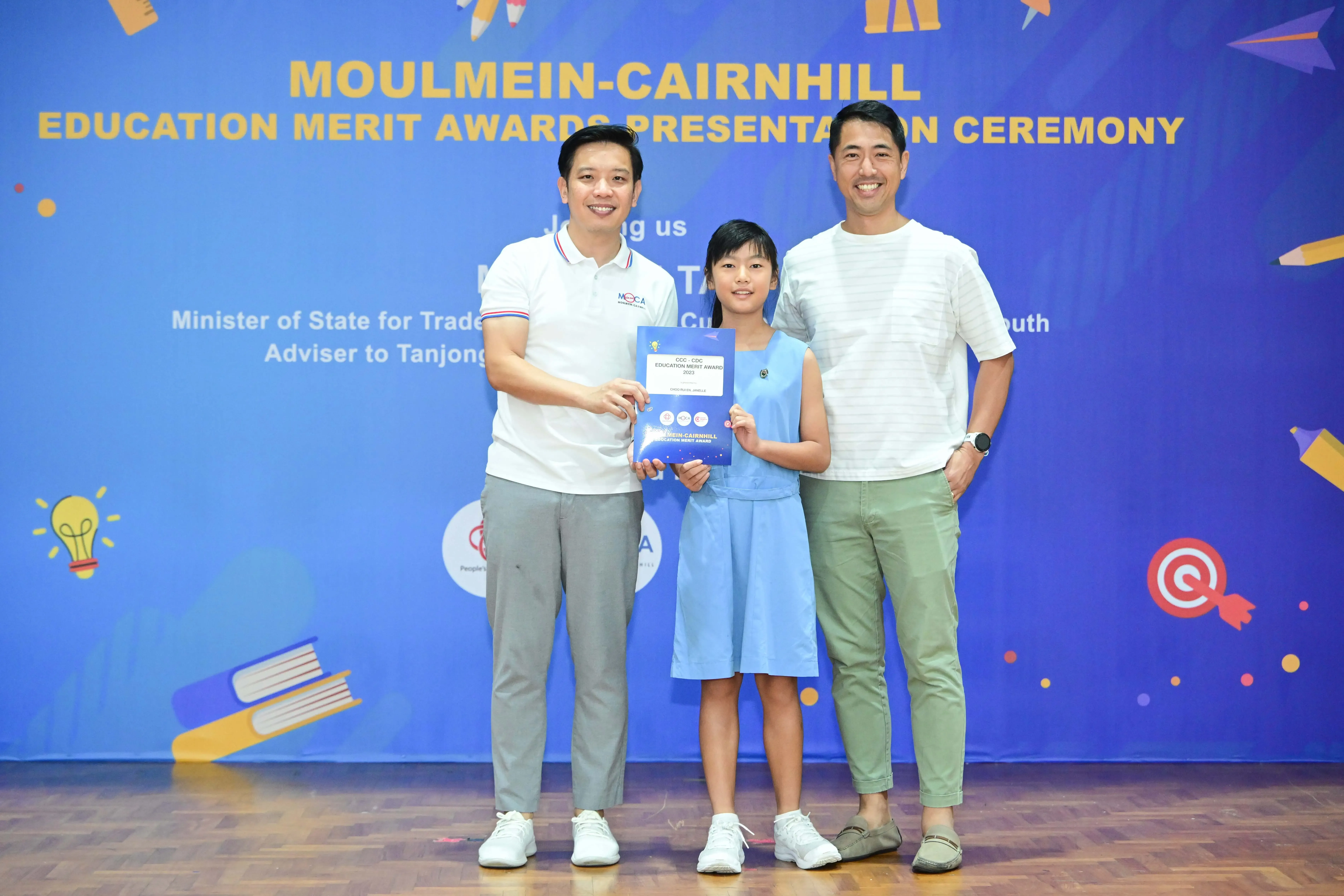 A girl holding an award certificate stands between two men at the Moulmein-Cairnhill Education Merit Awards ceremony.