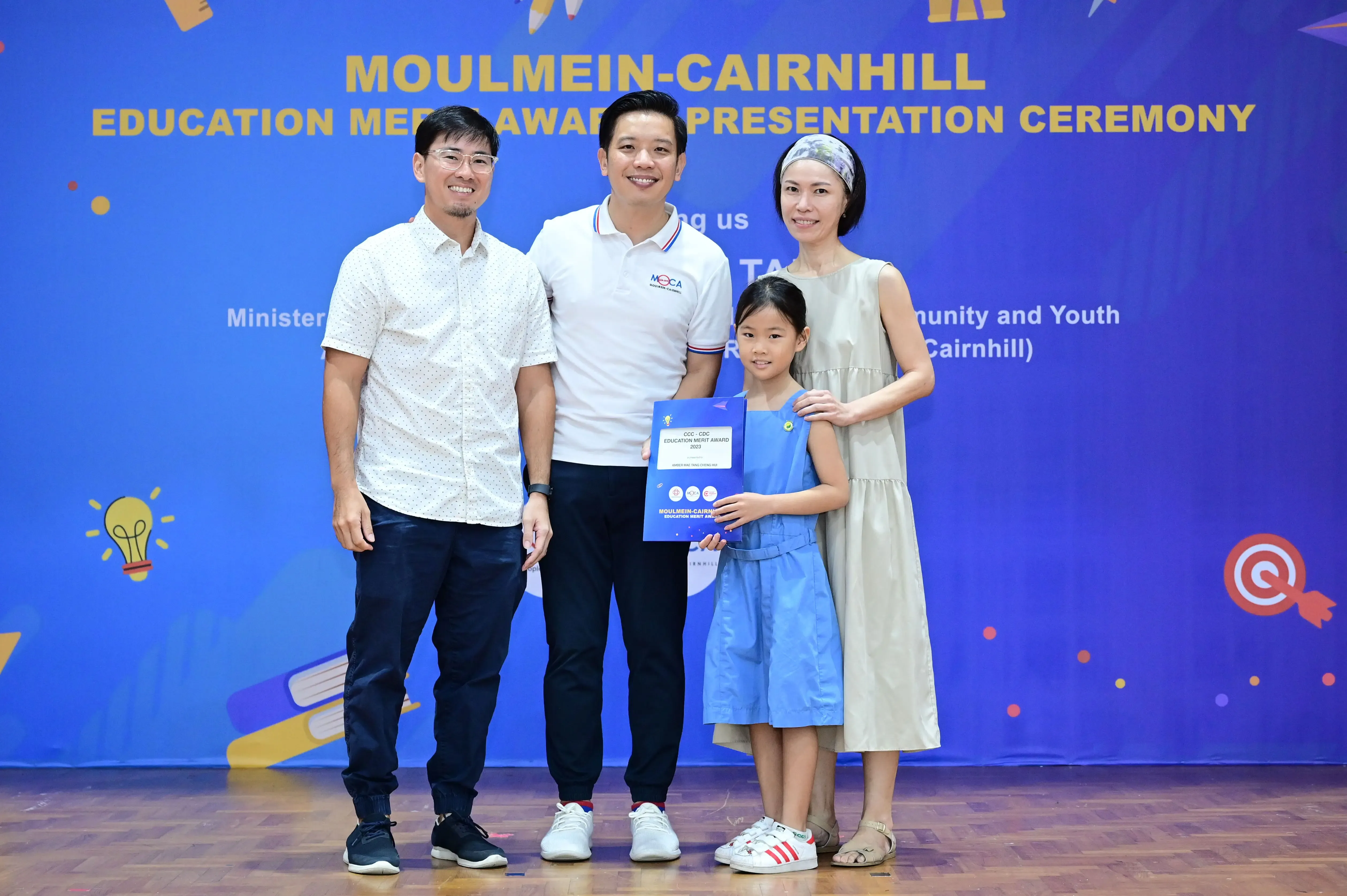Four people pose at the Moulmein-Cairnhill Education Merit Award Presentation Ceremony, with a girl holding a certificate.