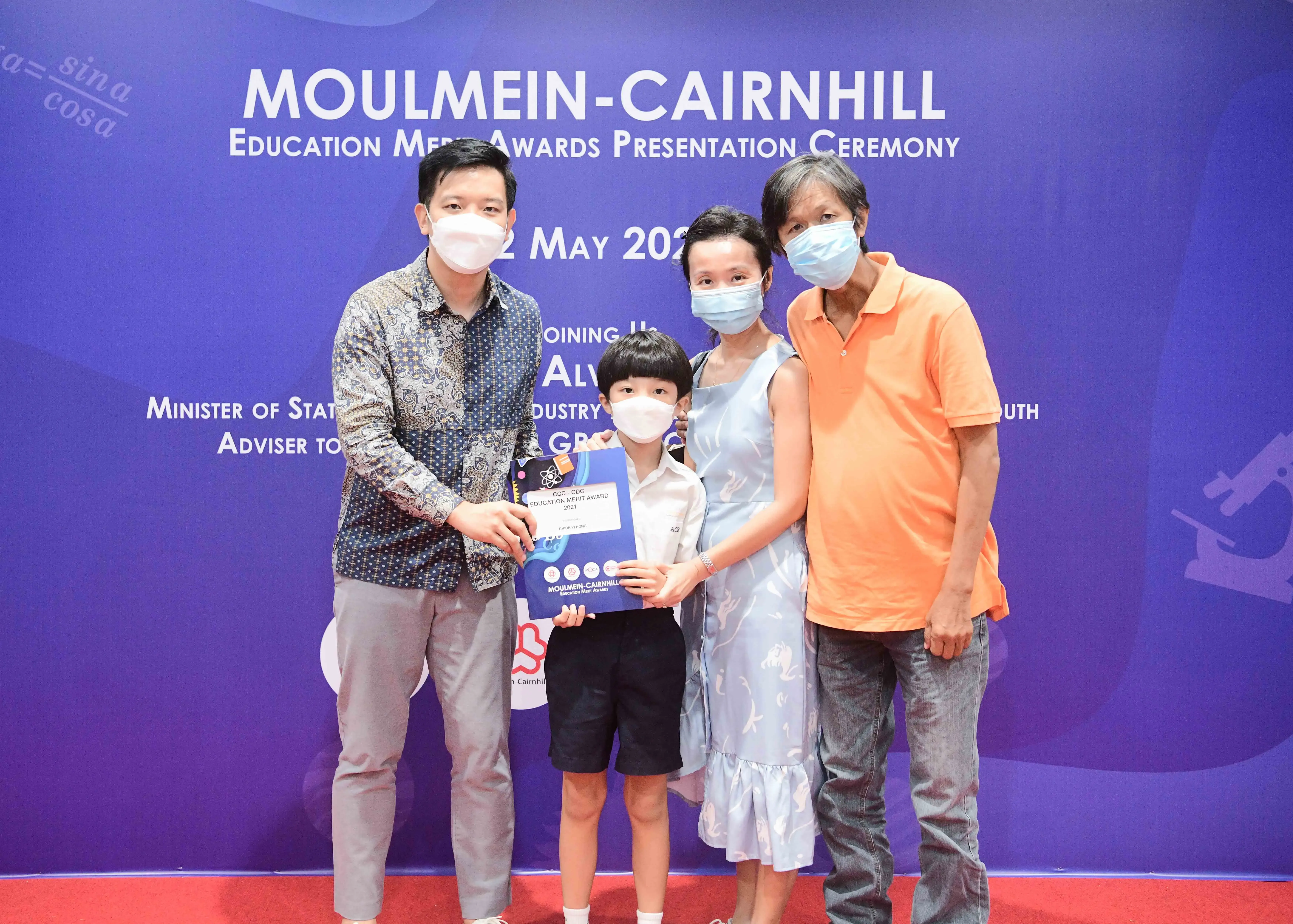 Four masked people pose at the Moulmein-Cairnhill Education Merit Awards Presentation Ceremony, with a boy holding a certificate.