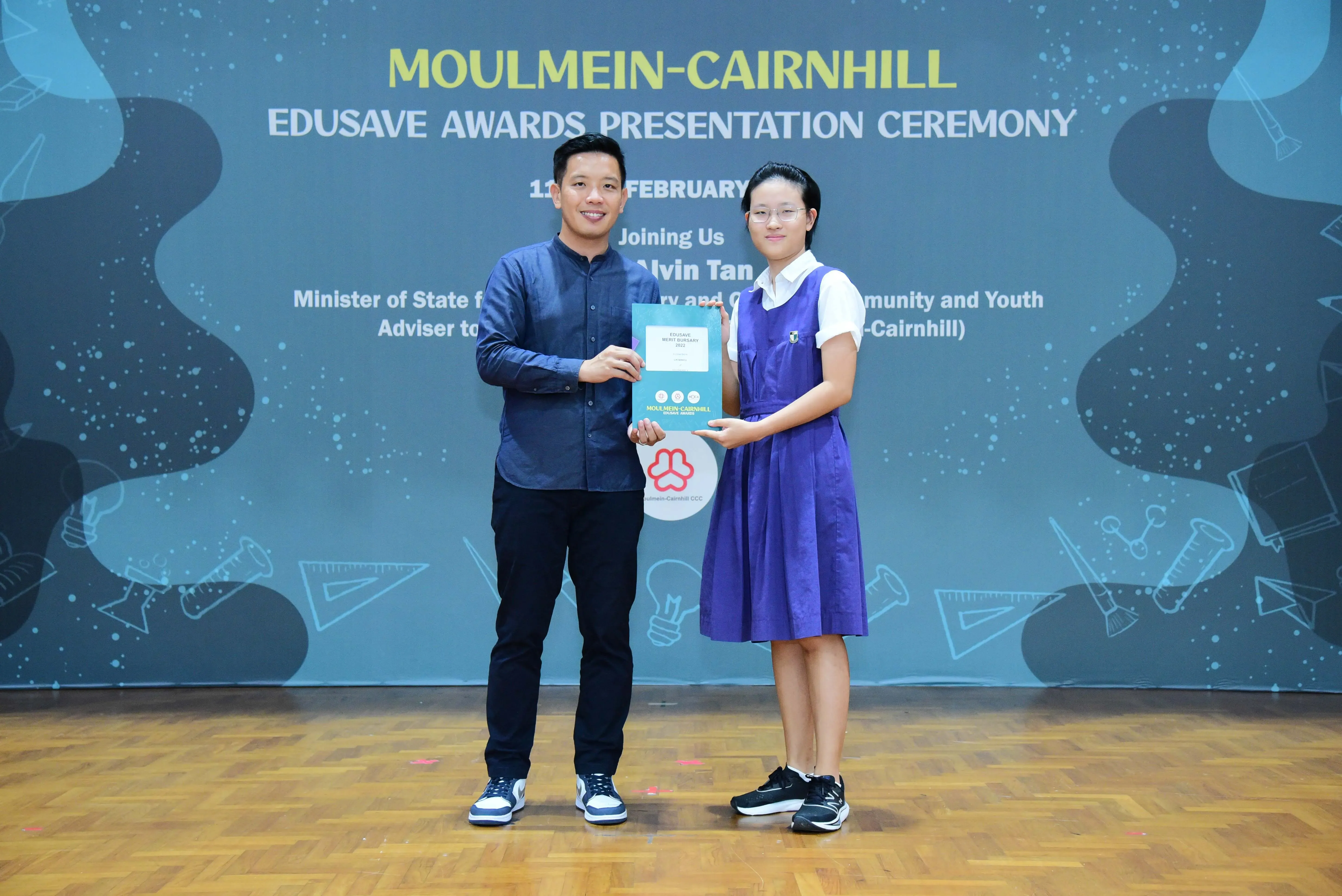 A man and a girl in school uniform hold a certificate together at the Moulmein-Cairnhill Edusave Awards Ceremony.
