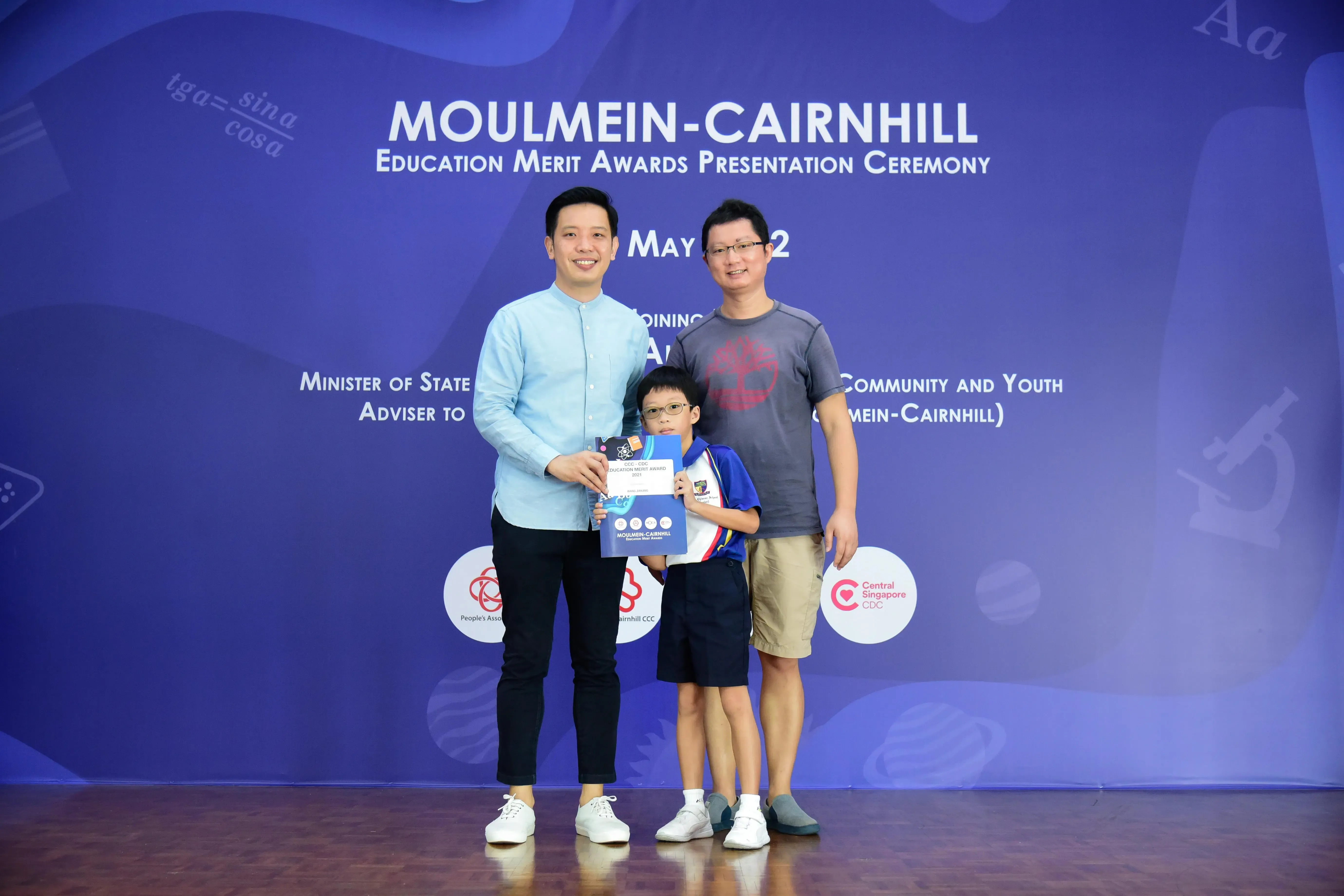 Two men and a young boy holding an award certificate stand before a Moulmein-Cairnhill Education Merit Awards backdrop.