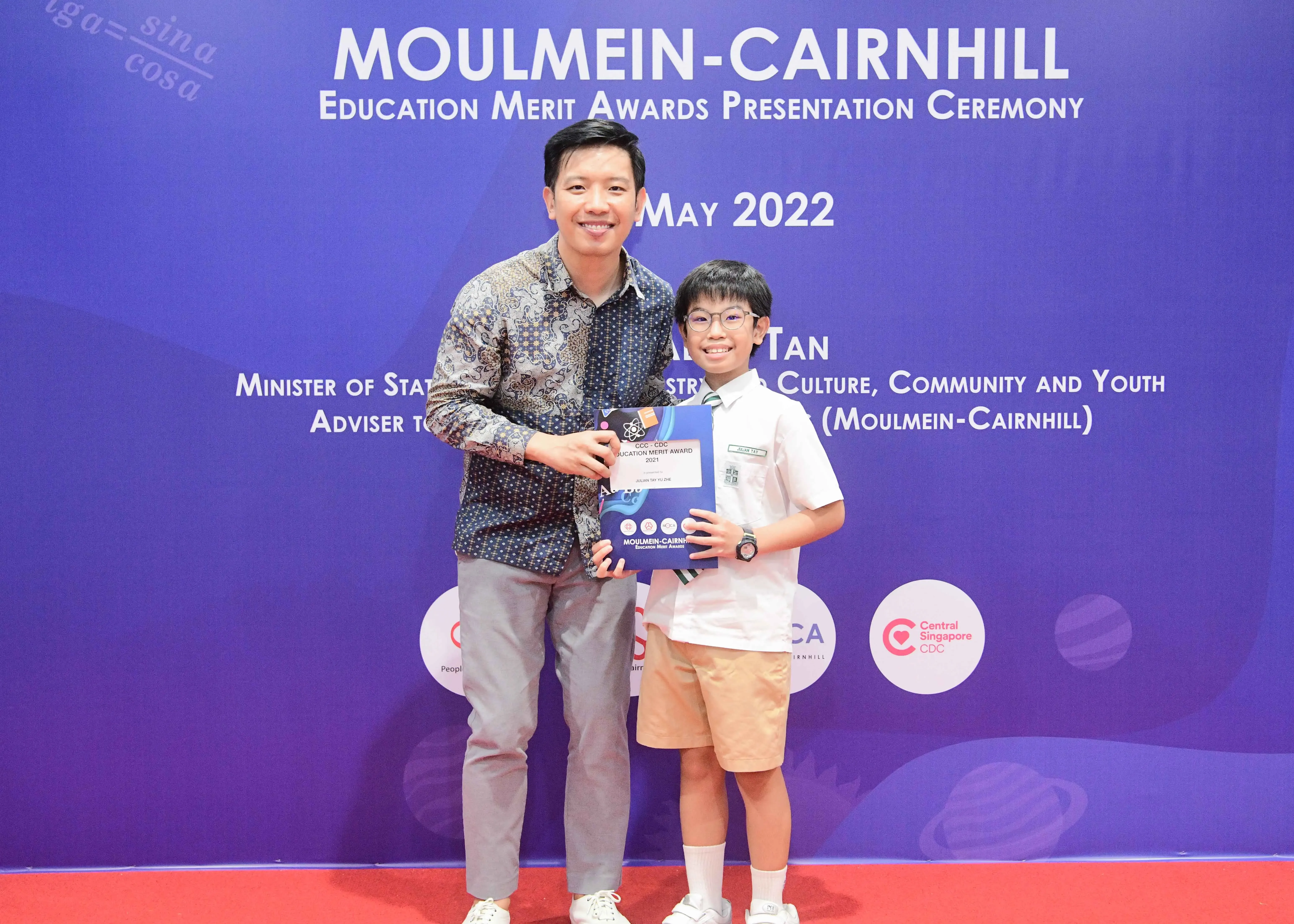 An adult and a young boy hold an award certificate at the Moulmein-Cairnhill Education Merit Awards Ceremony, May 2022.
