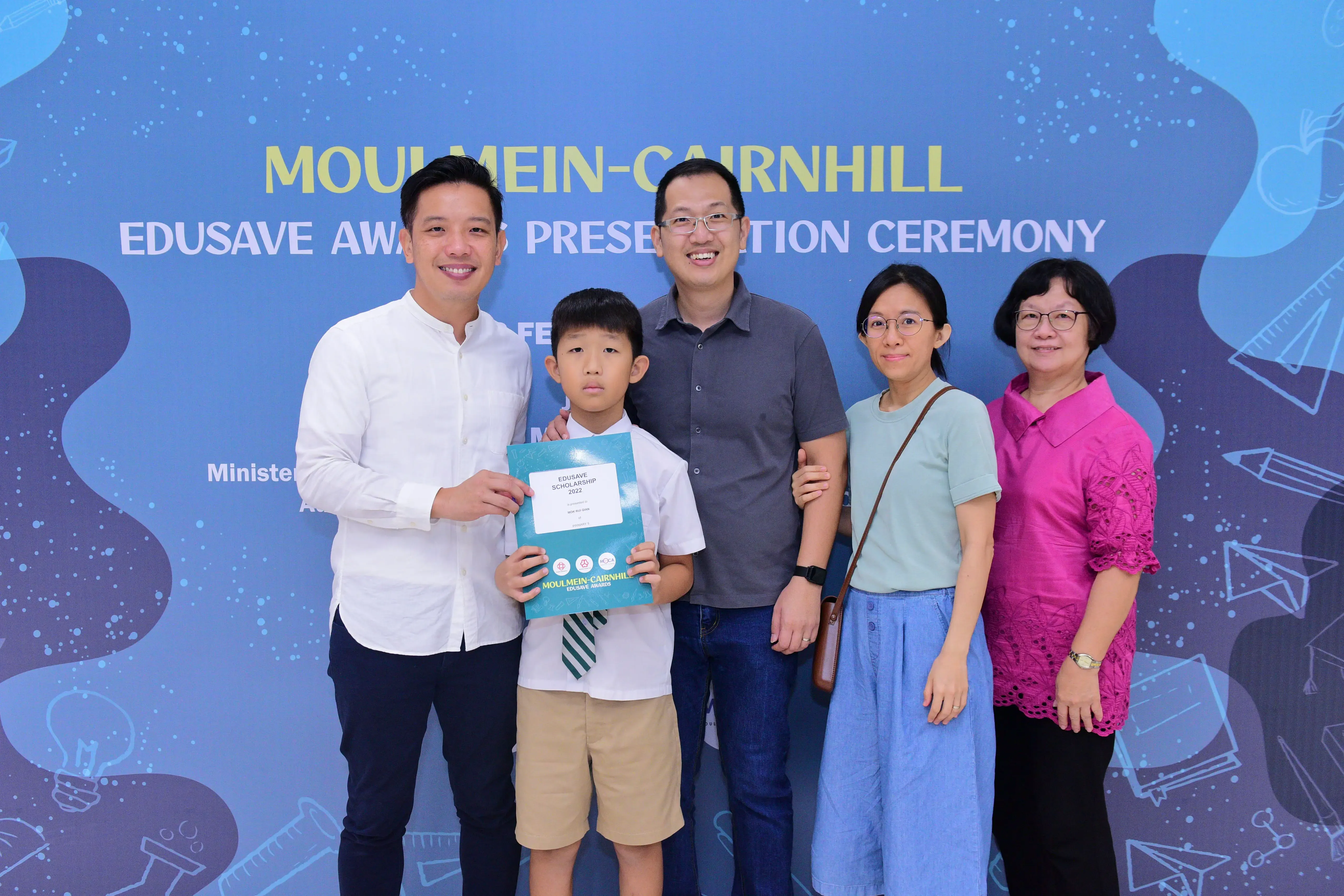 Five people pose at the Moulmein-Cairnhill Edusave Awards Ceremony, with a boy holding a scholarship certificate.