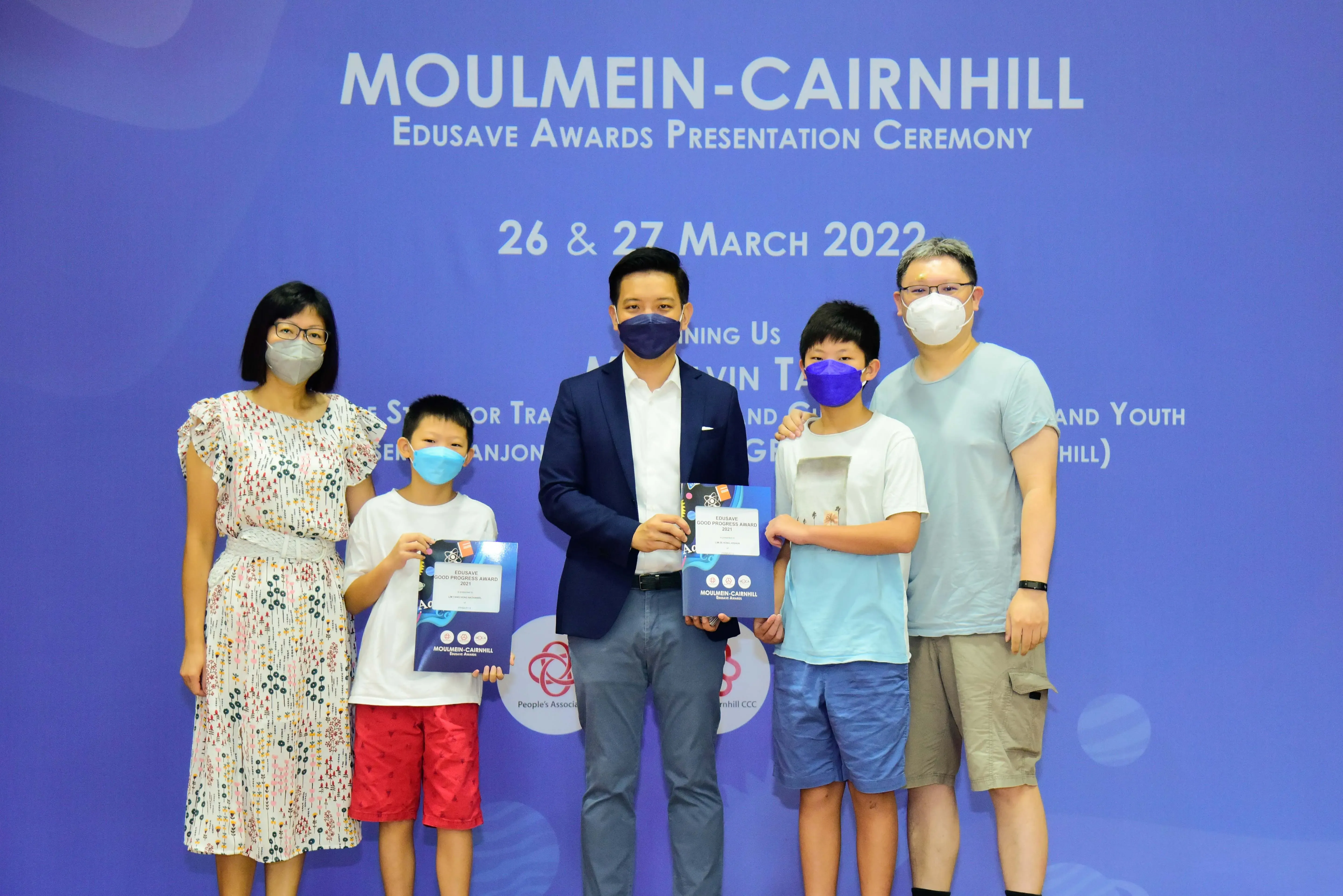 Five masked people pose at the Moulmein-Cairnhill Edusave Awards Ceremony; two children hold award folders.