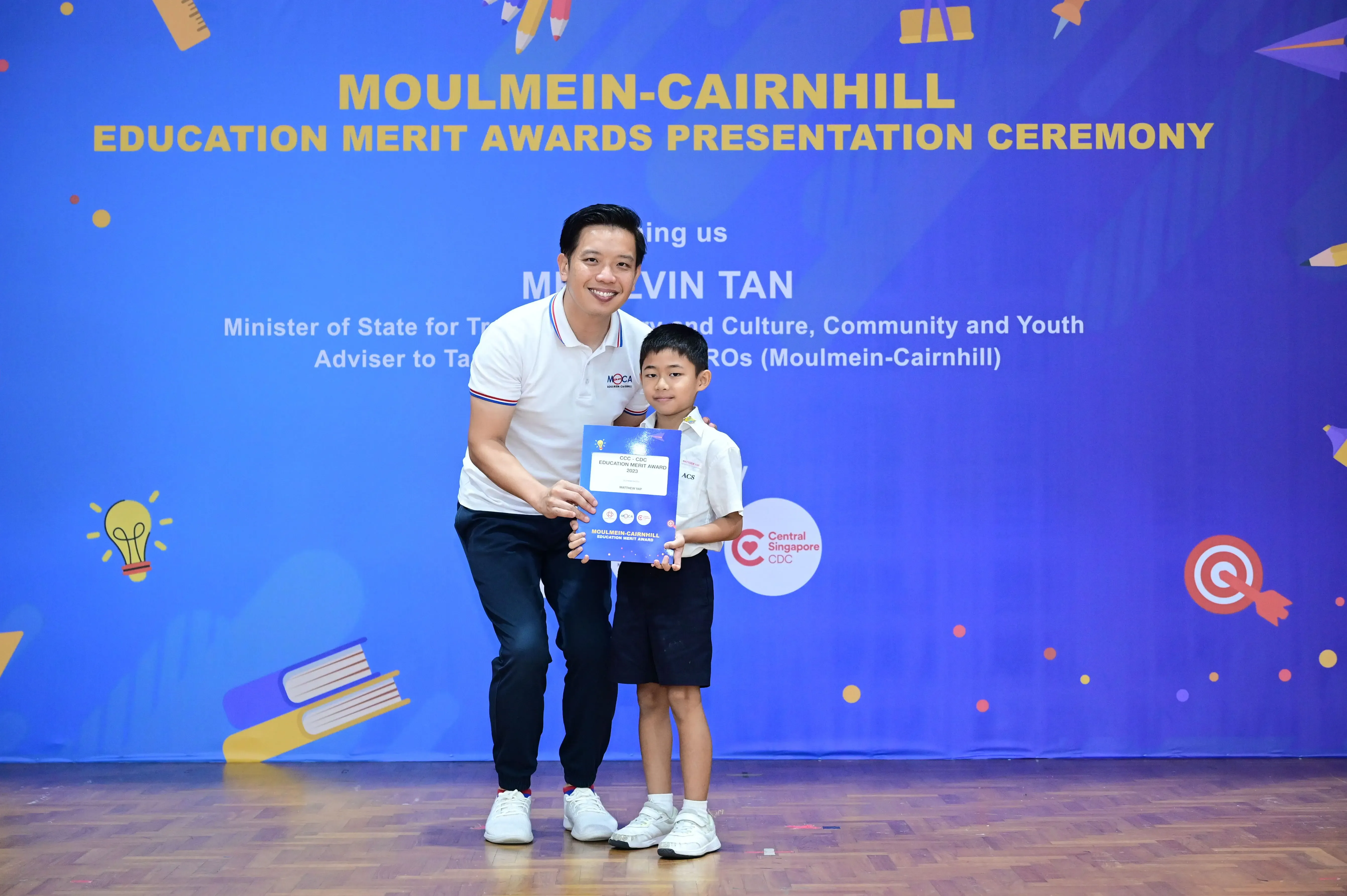 A man and a young boy hold a certificate together at the Moulmein-Cairnhill Education Merit Awards Presentation Ceremony.