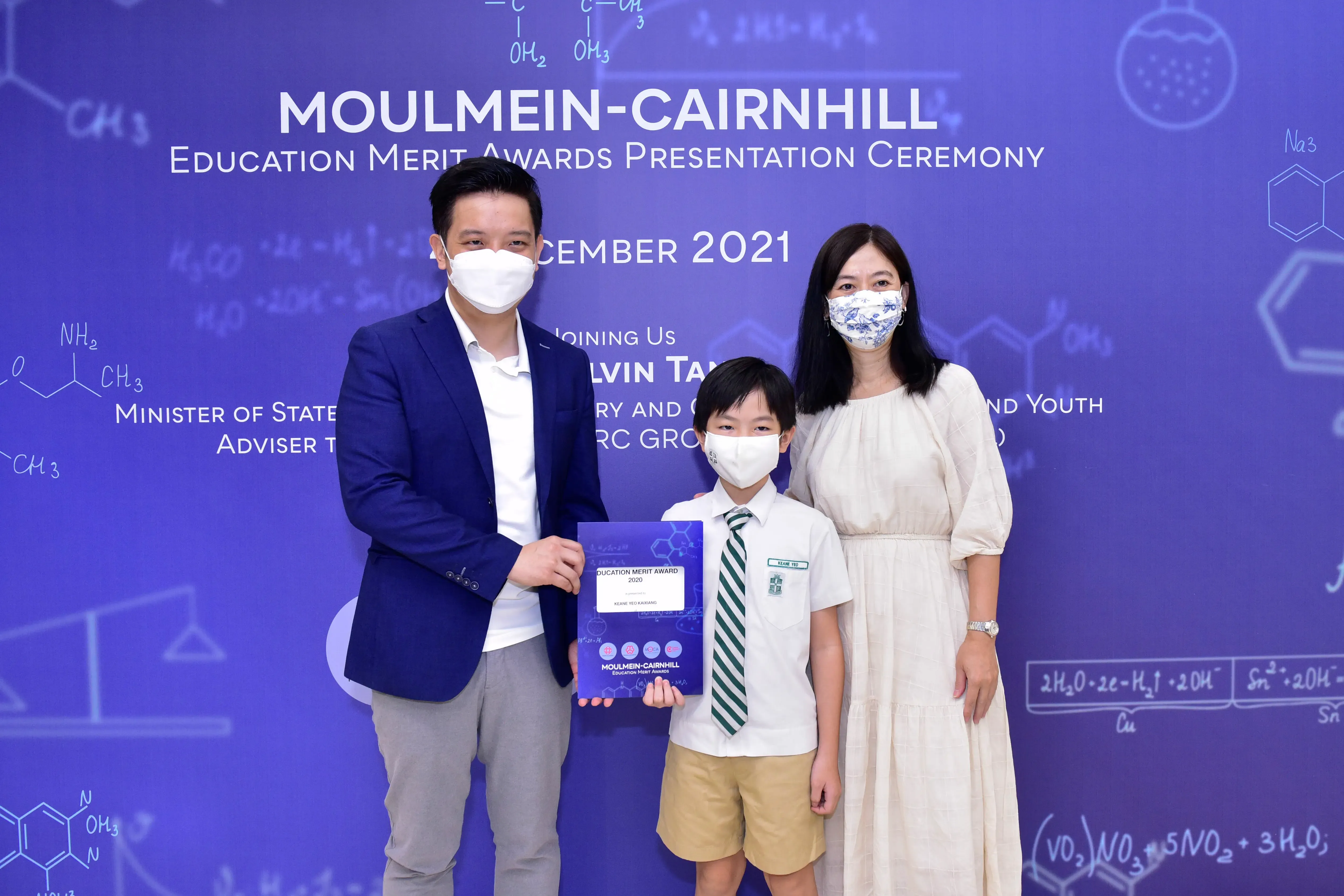 Two masked adults and a masked boy in school uniform hold an award at the Moulmein-Cairnhill Education Merit Awards 2021.
