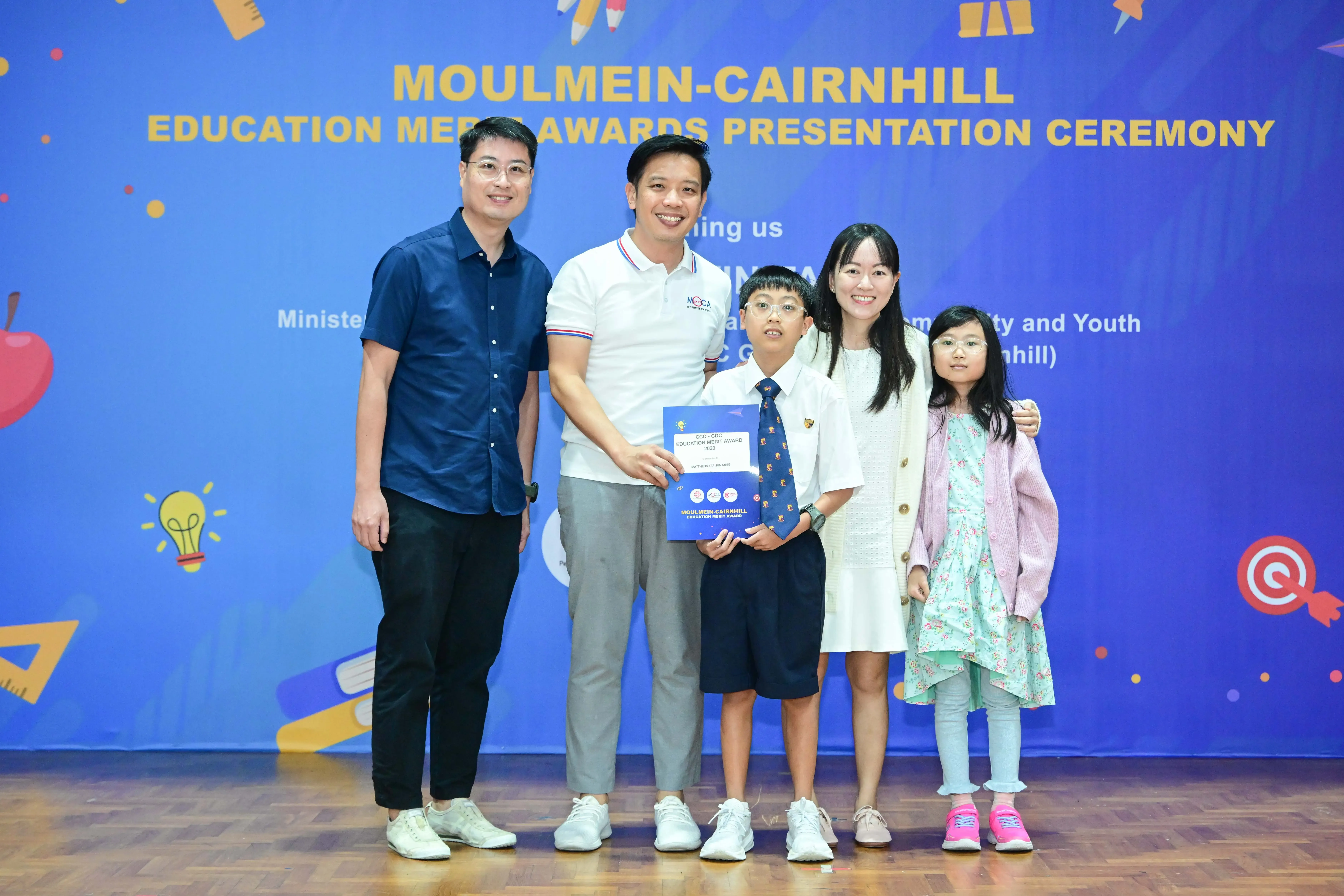 Five people pose in front of a Moulmein-Cairnhill Education Merit Awards Presentation Ceremony banner.