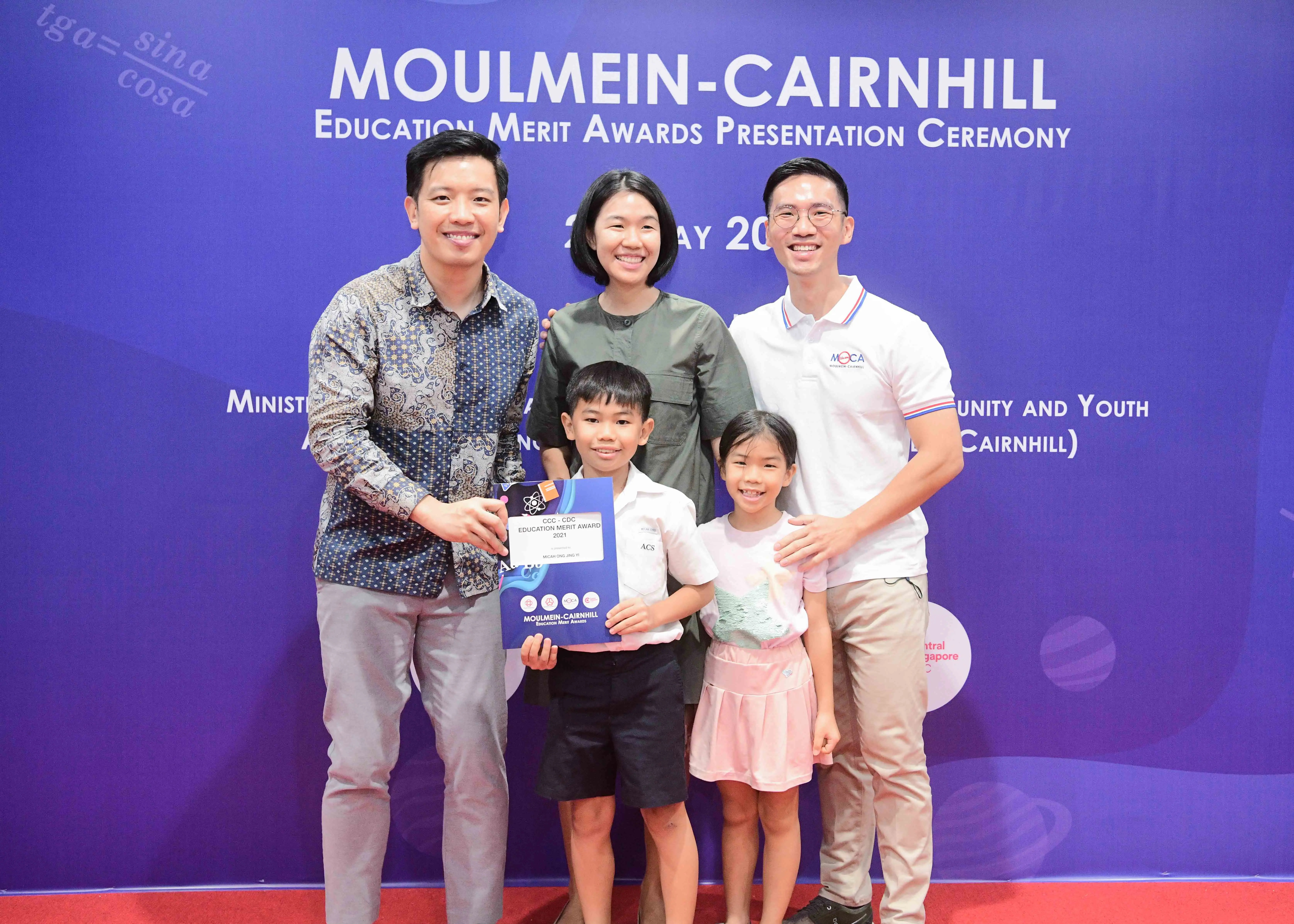 Five people smile at the Moulmein-Cairnhill Education Merit Awards ceremony, with a boy holding an award certificate.