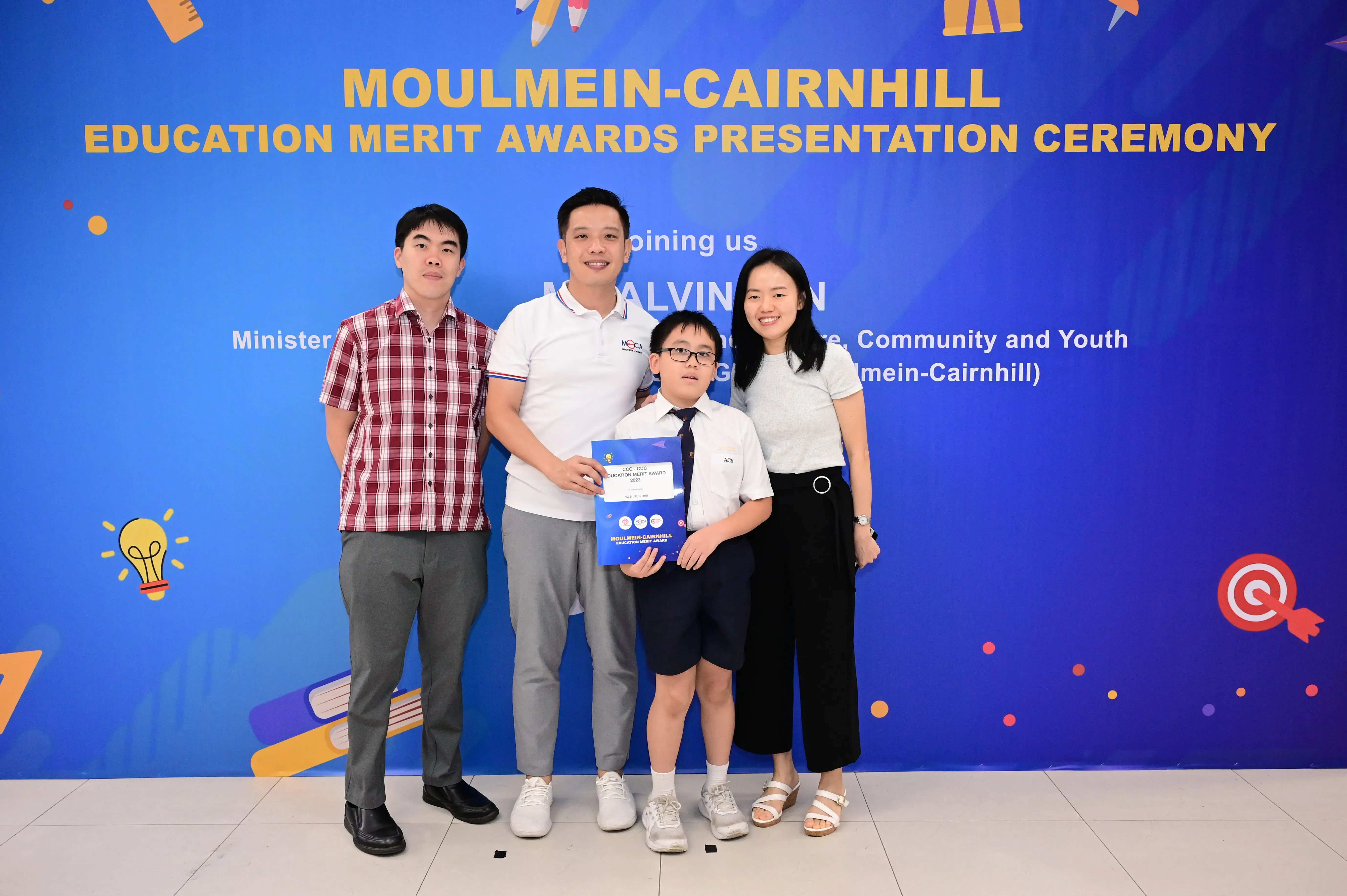 Three adults and a boy holding an award folder pose before a Moulmein-Cairnhill Education Merit Awards banner.