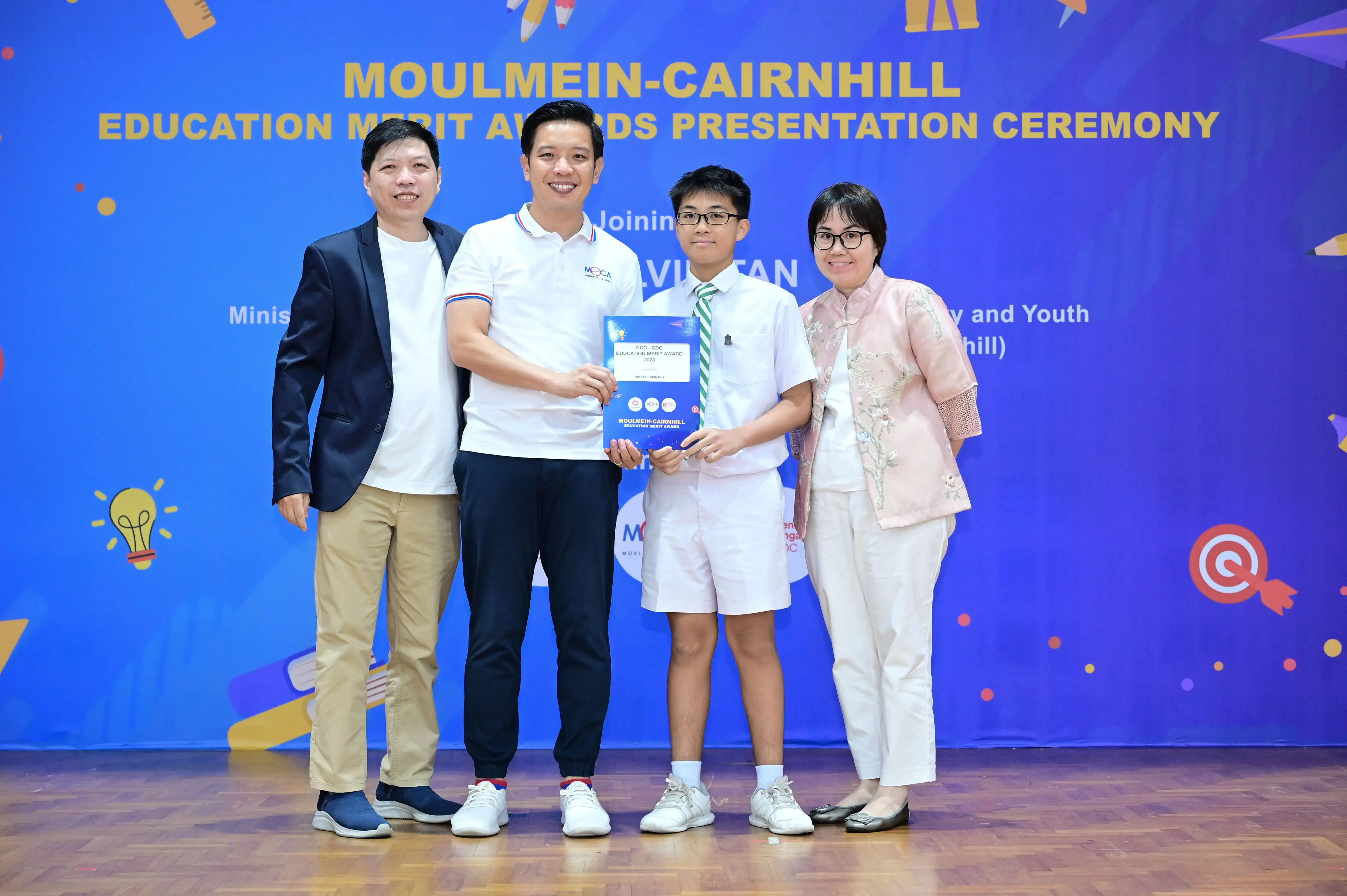 Four people pose at the Moulmein-Cairnhill Education Merit Awards Presentation Ceremony, with a boy holding an award certificate.