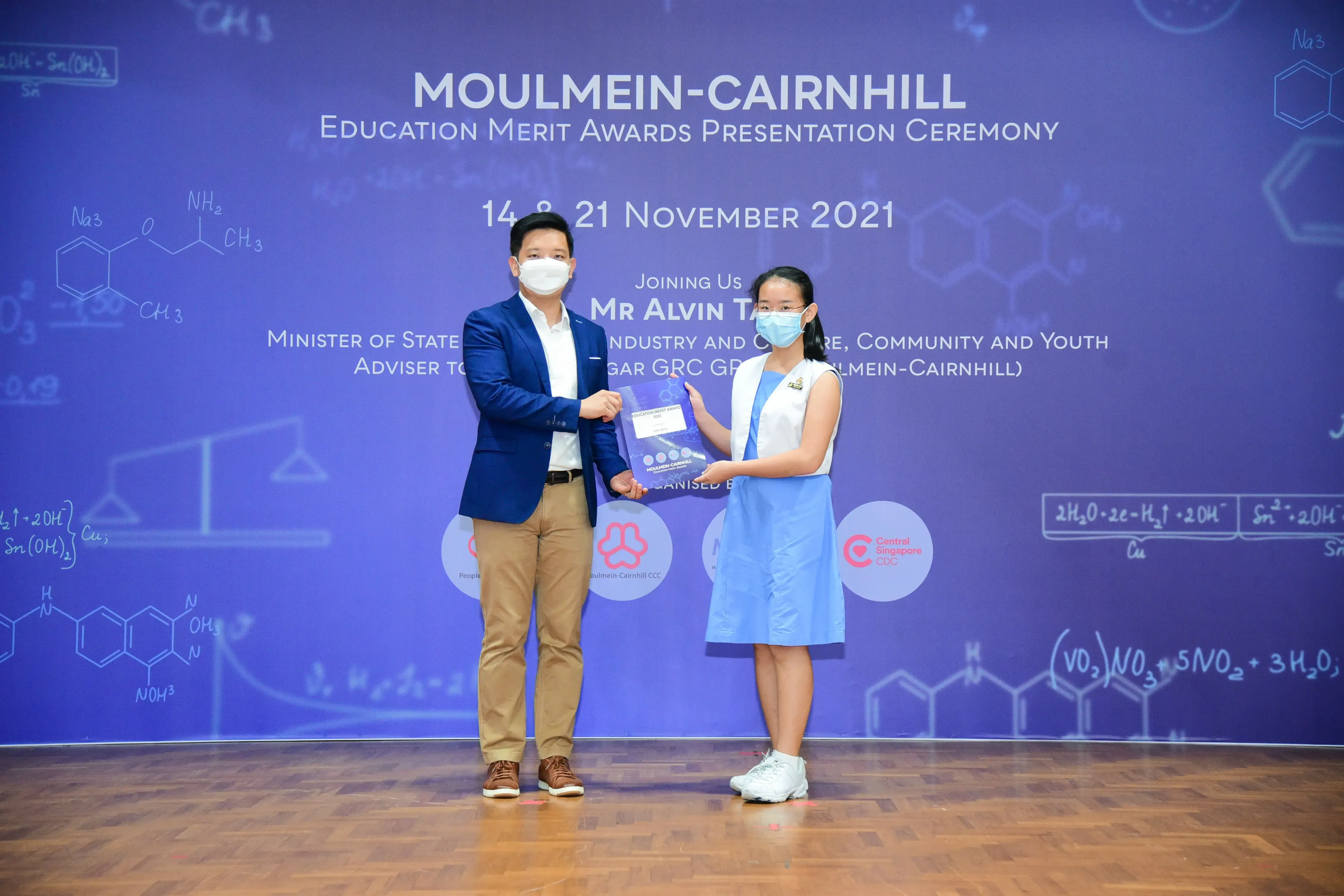 A man in a blue blazer presents an award to a young girl at the Moulmein-Cairnhill Education Merit Awards Ceremony 2021.