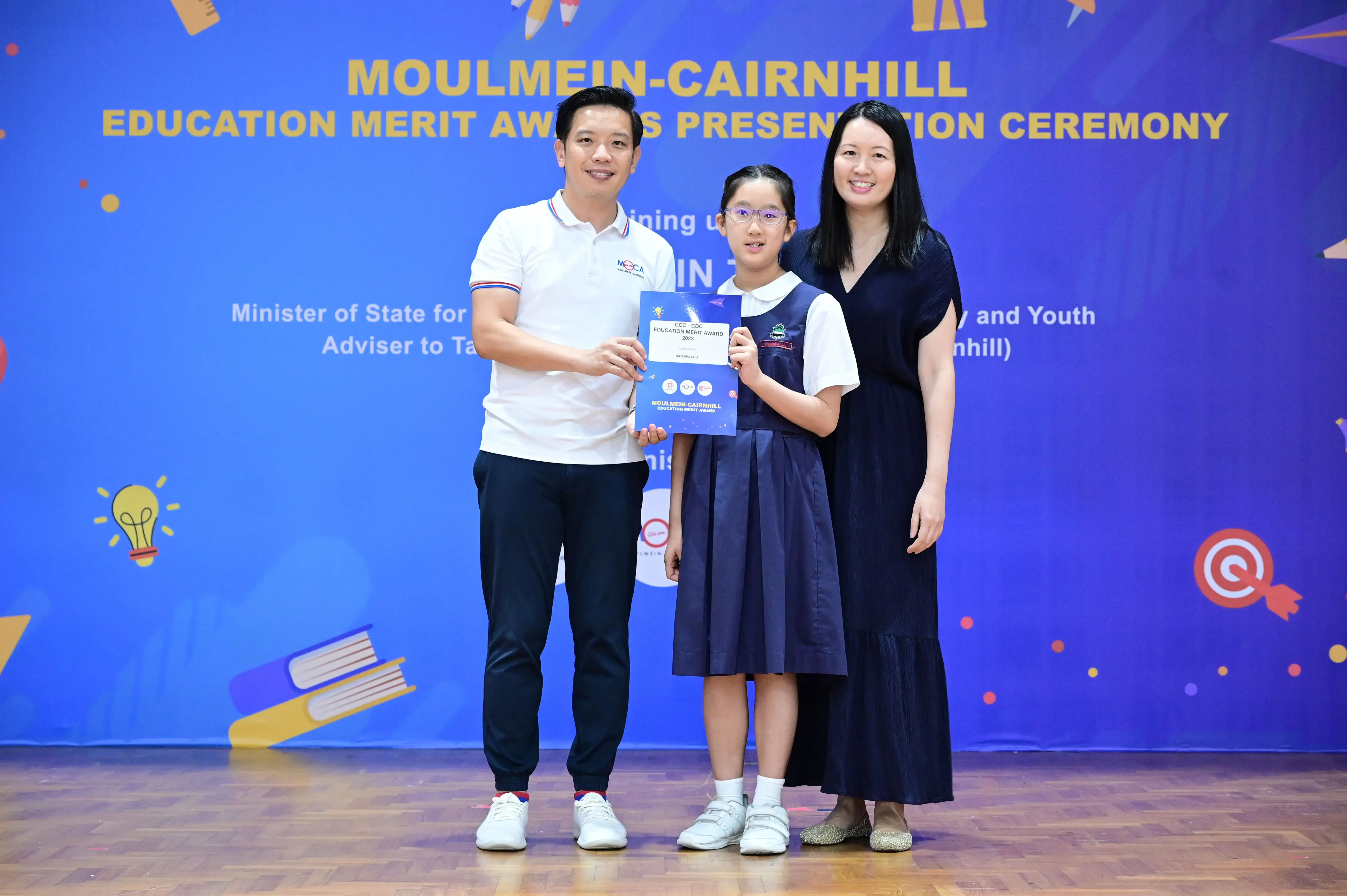 A girl in school uniform holds a merit award certificate flanked by two adults at the Moulmein-Cairnhill Education Merit Awards ceremony.