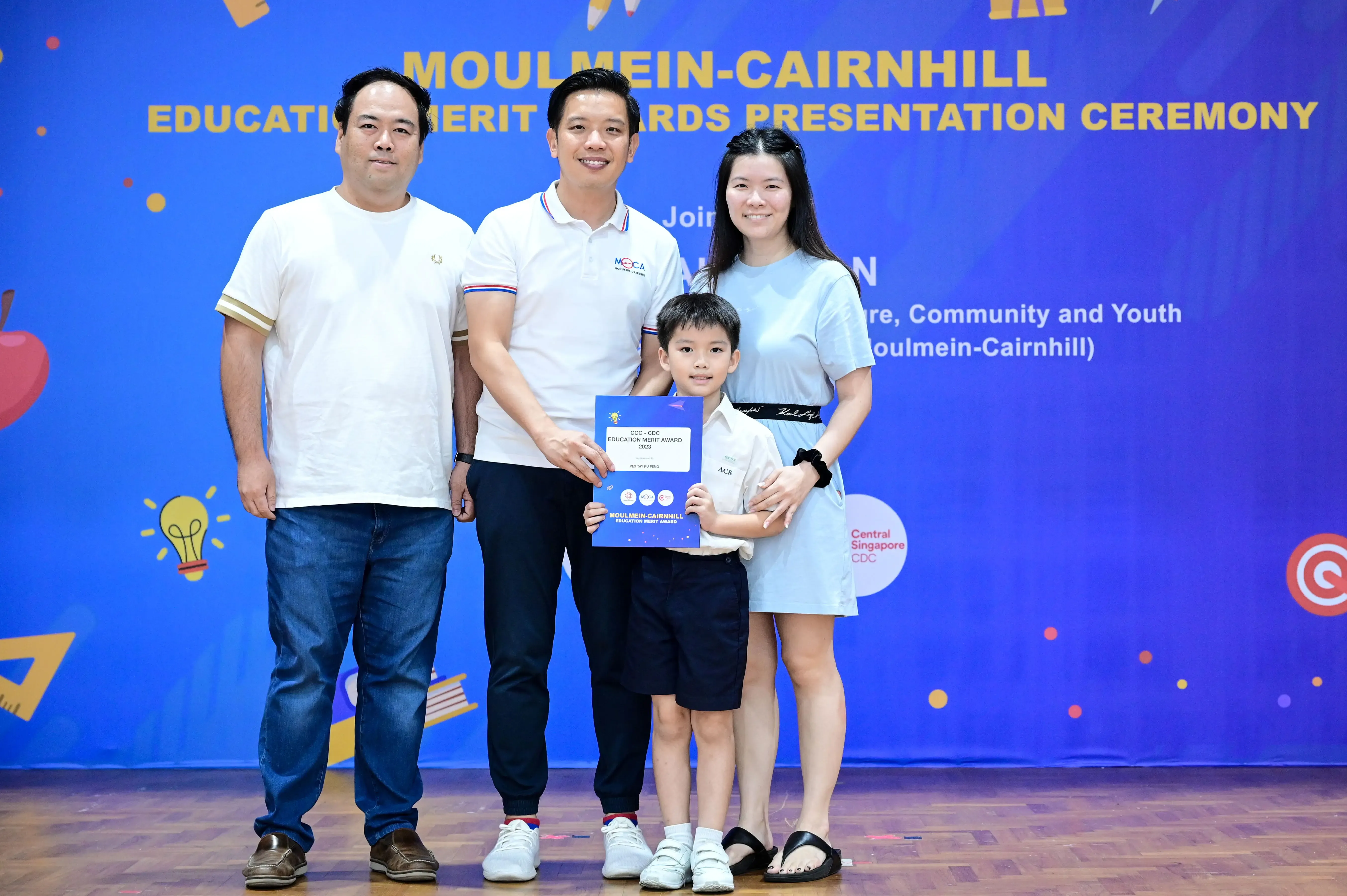 Three adults and a young boy pose holding a blue award certificate at the Moulmein-Cairnhill Education Merit Awards ceremony.