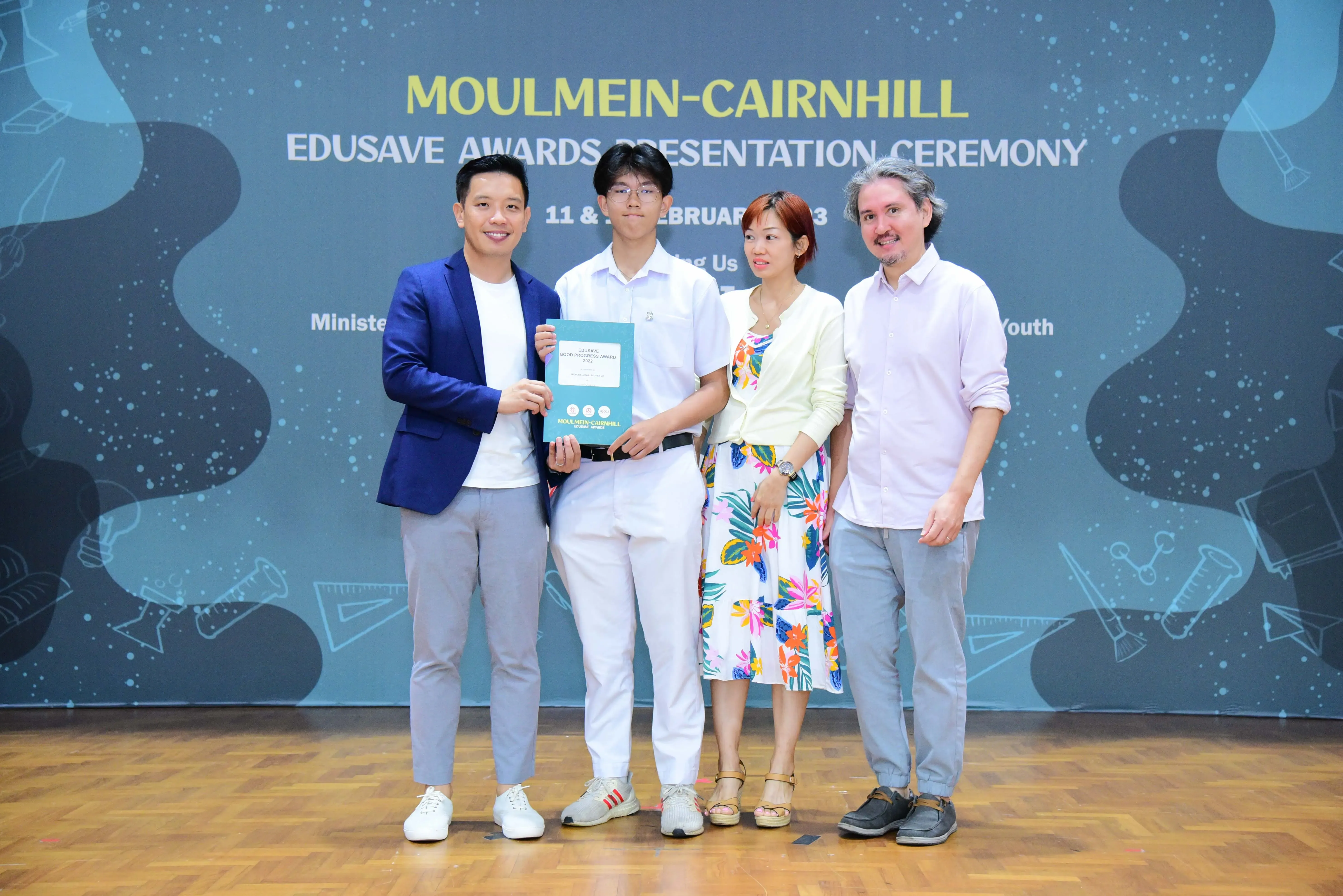 Four people pose with a certificate at the Moulmein-Cairnhill Edusave Awards Presentation Ceremony backdrop.