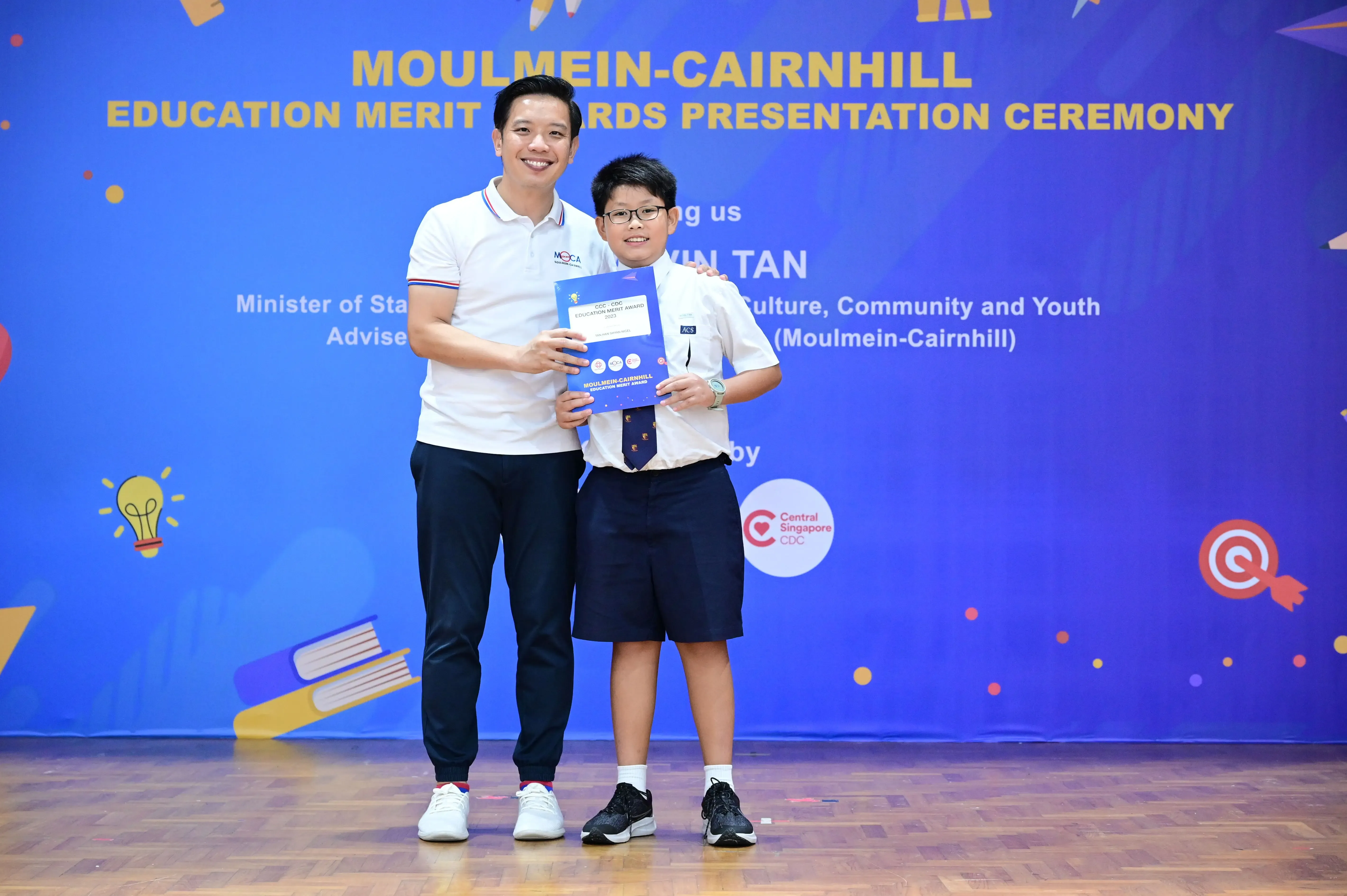 An adult and a young student hold a certificate at the Moulmein-Cairnhill Education Merit Awards Presentation Ceremony.