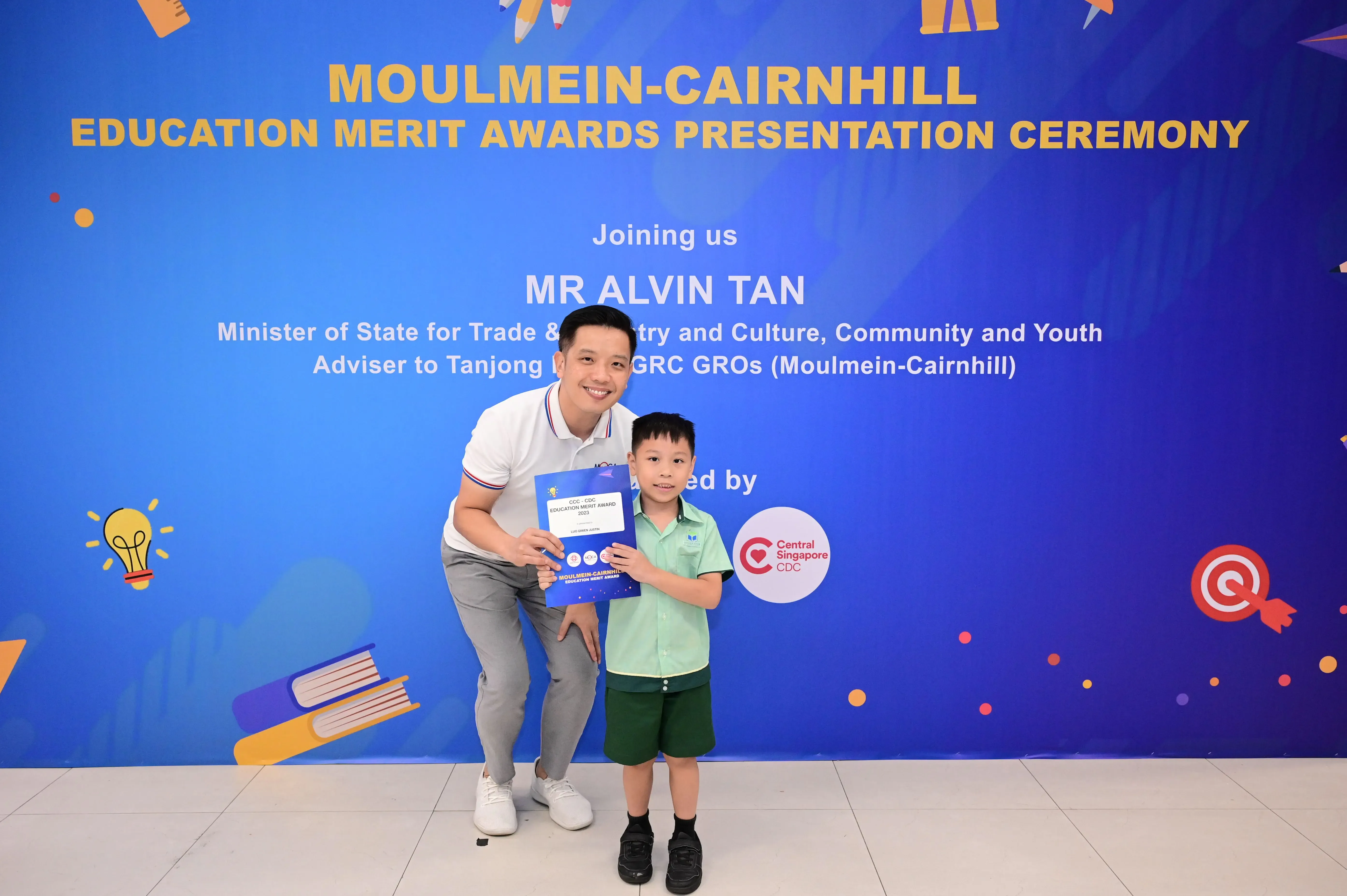 Man and young boy hold a blue award certificate in front of a Moulmein-Cairnhill Education Merit Awards banner.