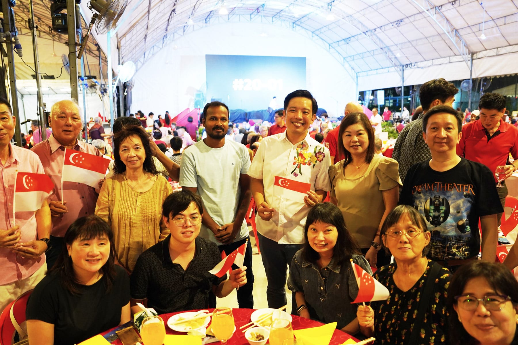 Group of smiling people holding small Singapore flags at a festive indoor community event under a tent.