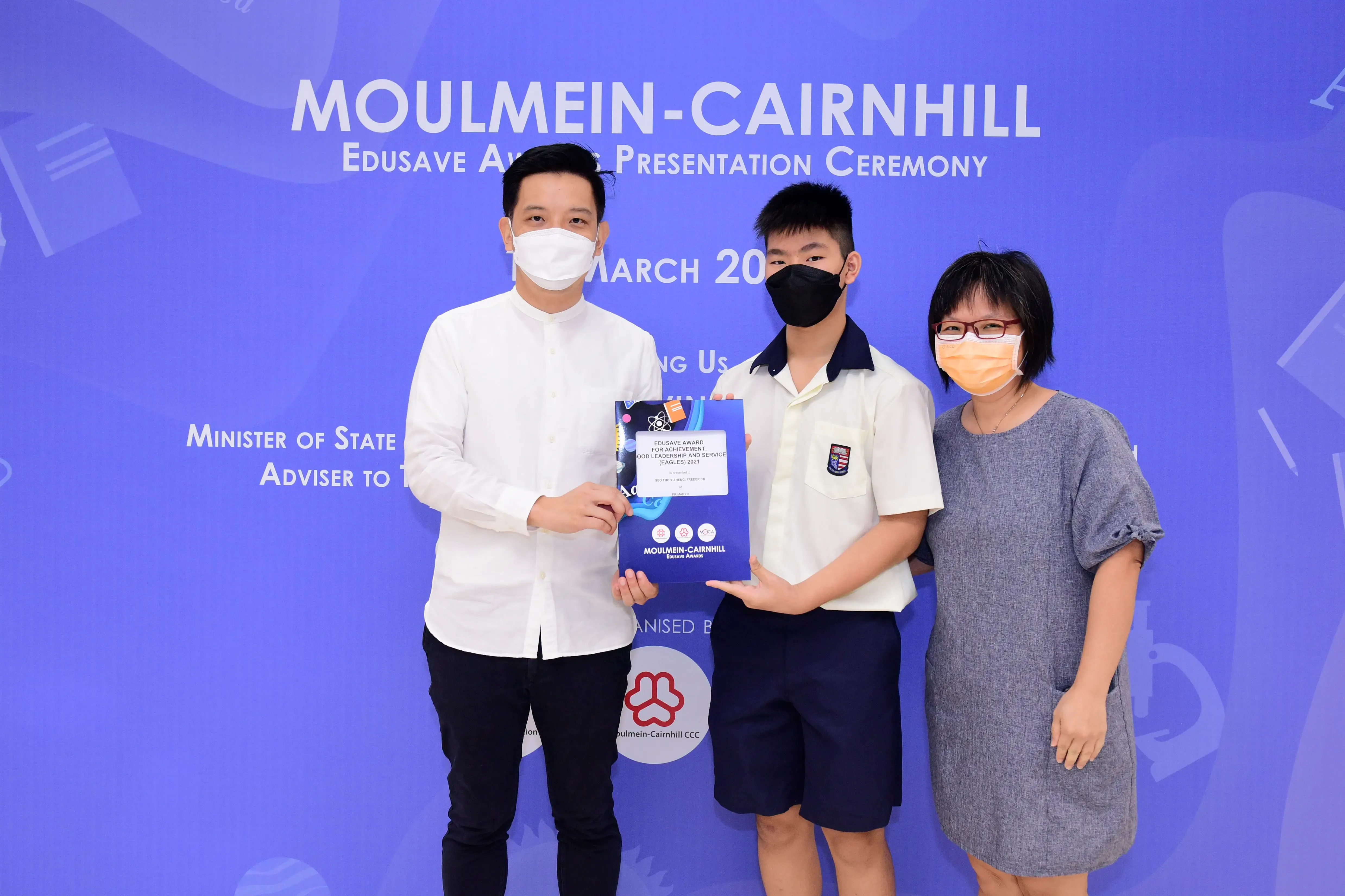 Three masked people pose at the Moulmein-Cairnhill Edusave Award Presentation Ceremony, with a student holding an award folder.