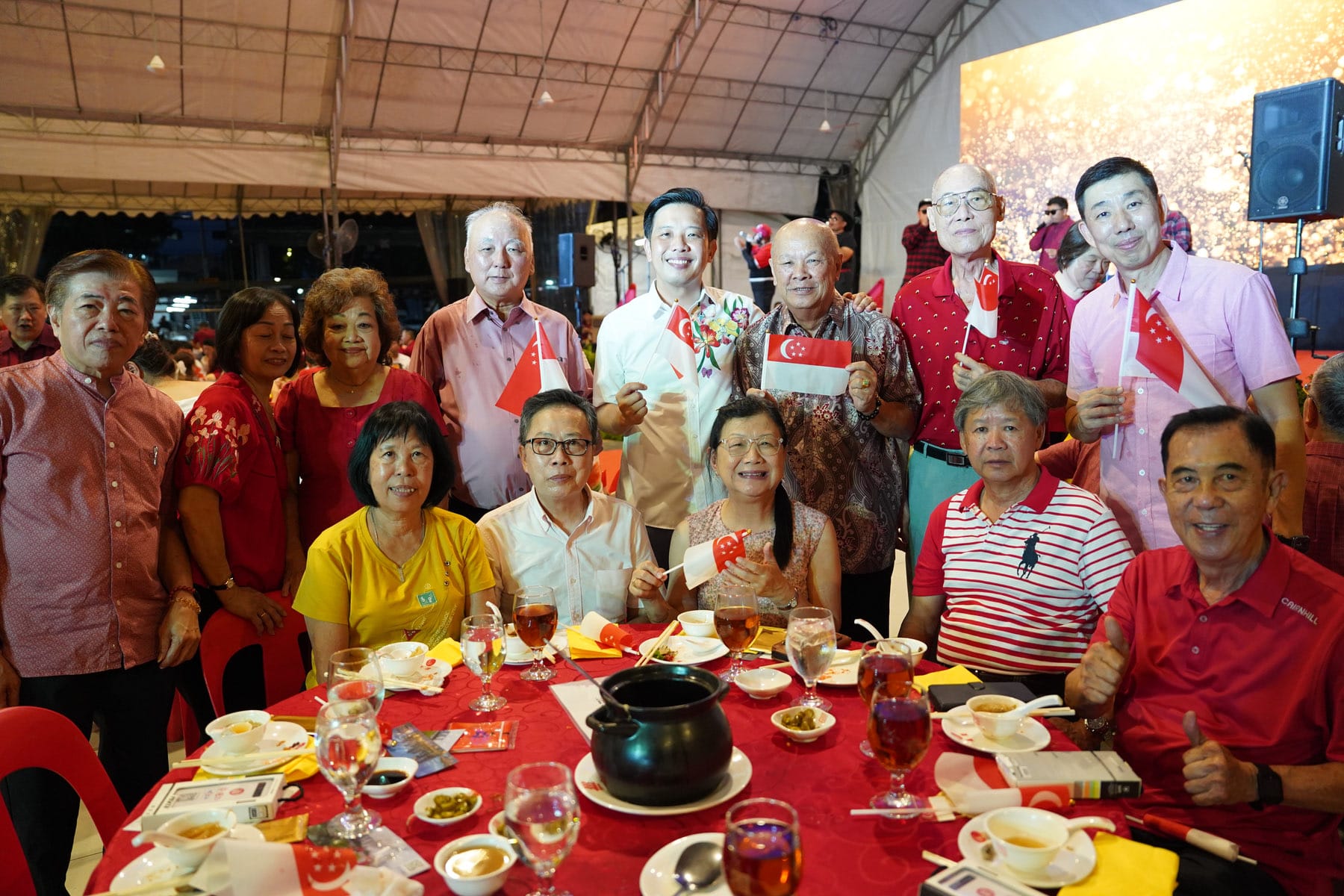 Group of people in red outfits posing at a banquet table, holding small Singapore flags at an outdoor celebration event.