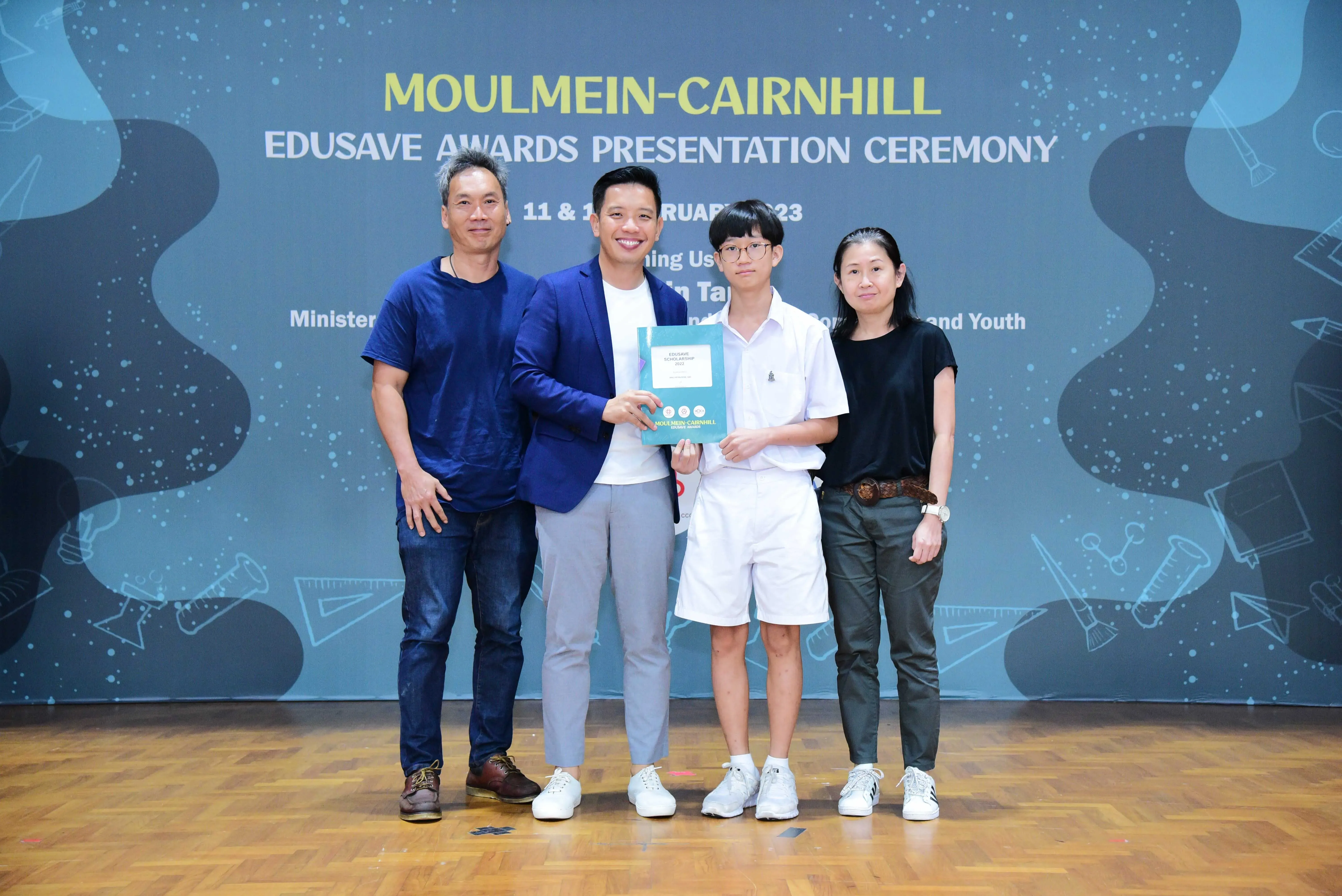 Four people pose at the Moulmein-Cairnhill Edusave Awards Presentation Ceremony, with a student holding an award booklet.
