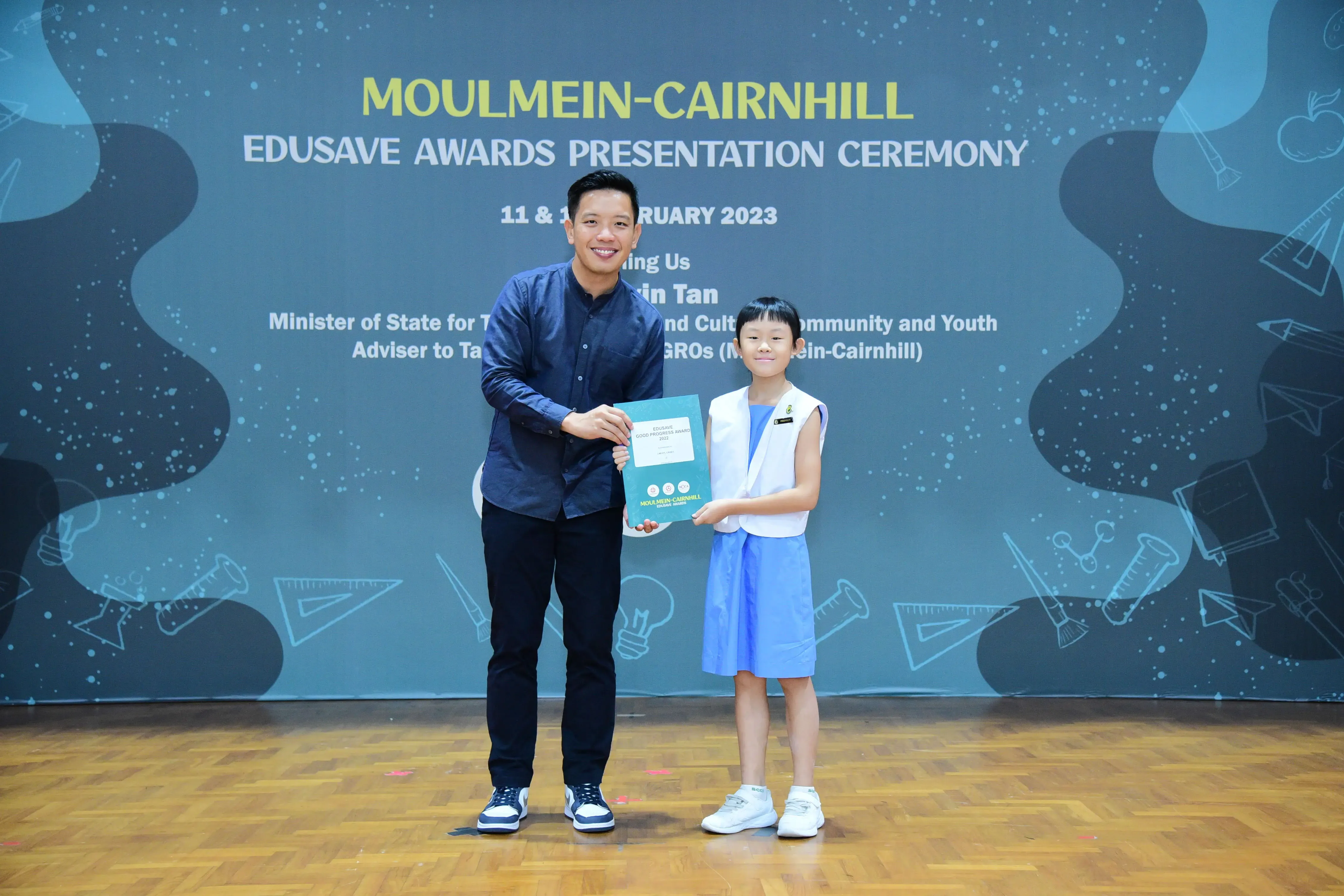 A man and a young girl hold a certificate together on stage at the Moulmein-Cairnhill Edusave Awards Ceremony 2023.