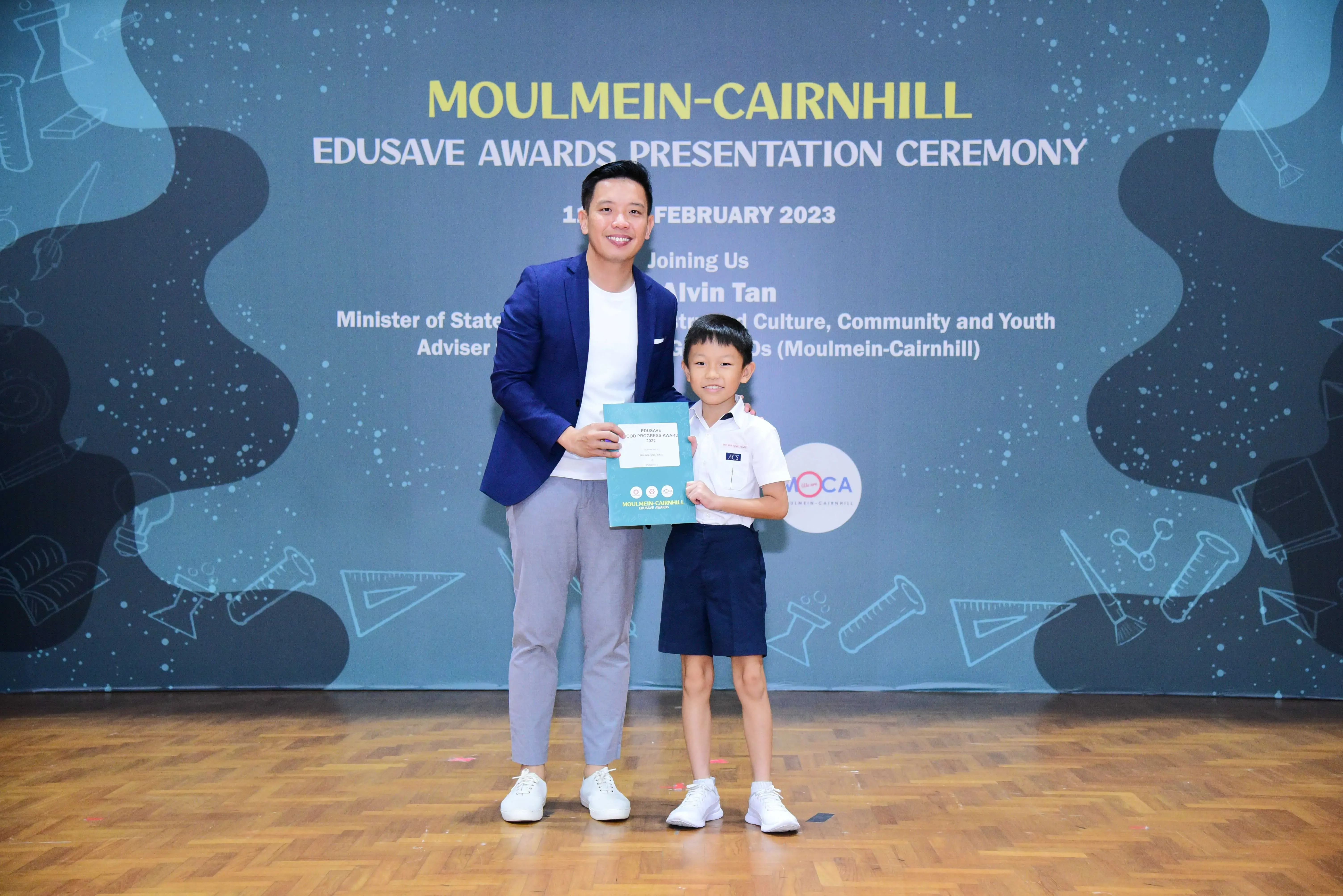 A man in a blue blazer and a young boy in school uniform hold a certificate at the Moulmein-Cairnhill Edusave Awards 2023.