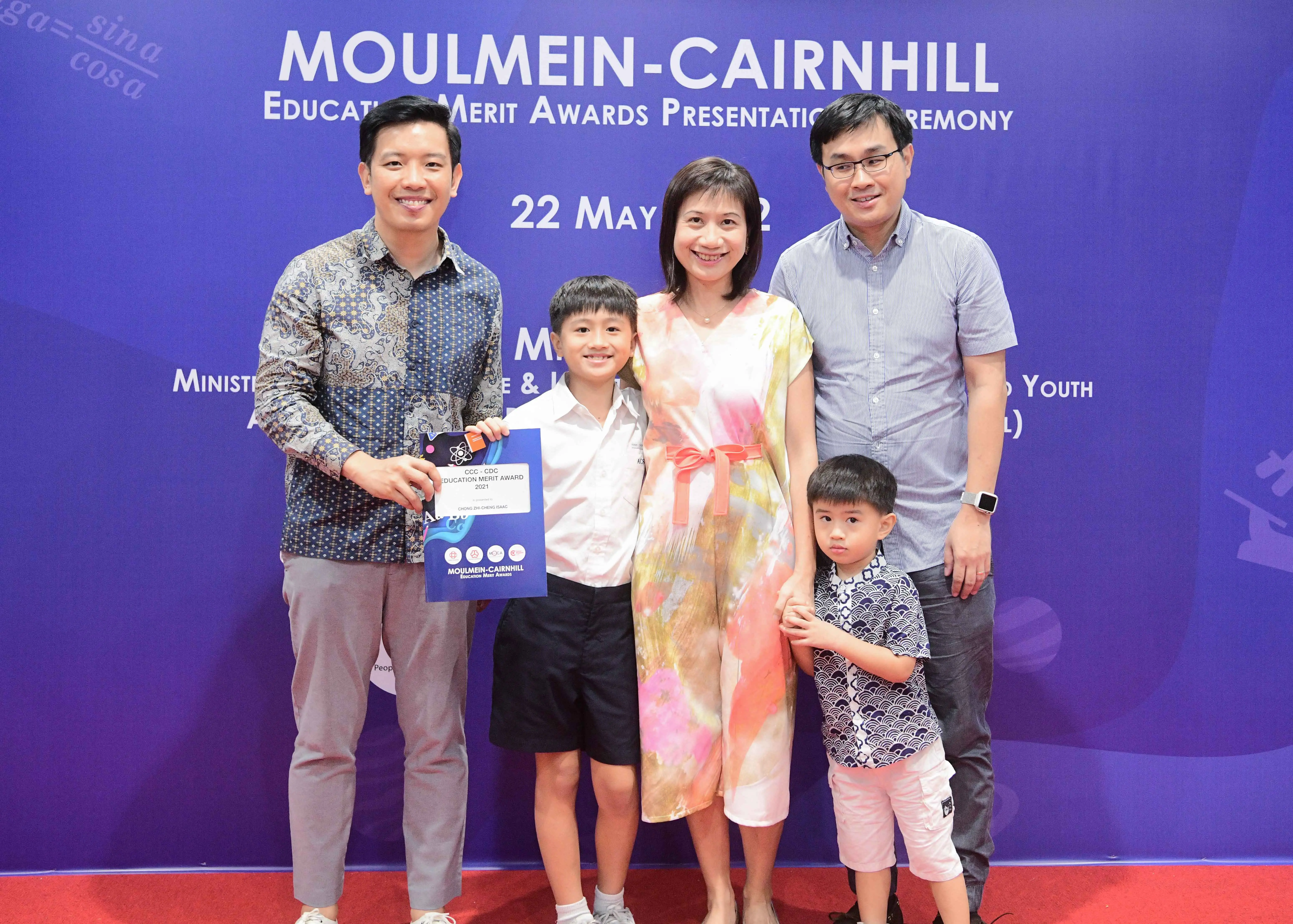 Five people pose at the Moulmein-Cairnhill Education Merit Awards Presentation Ceremony, including two children holding a certificate.