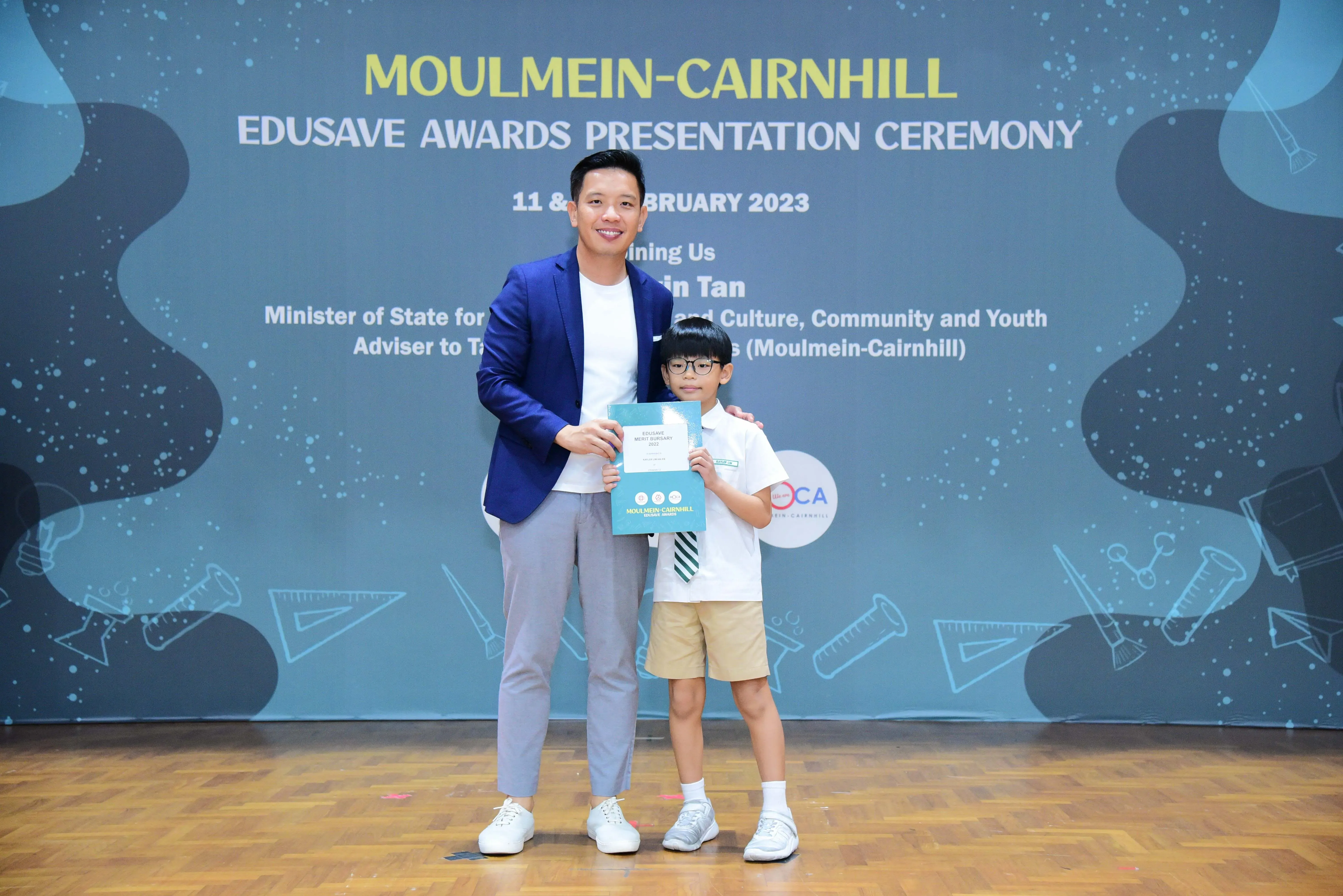 A man in a blue blazer and a young boy in school uniform hold a certificate at the Moulmein-Cairnhill Edusave Awards 2023.