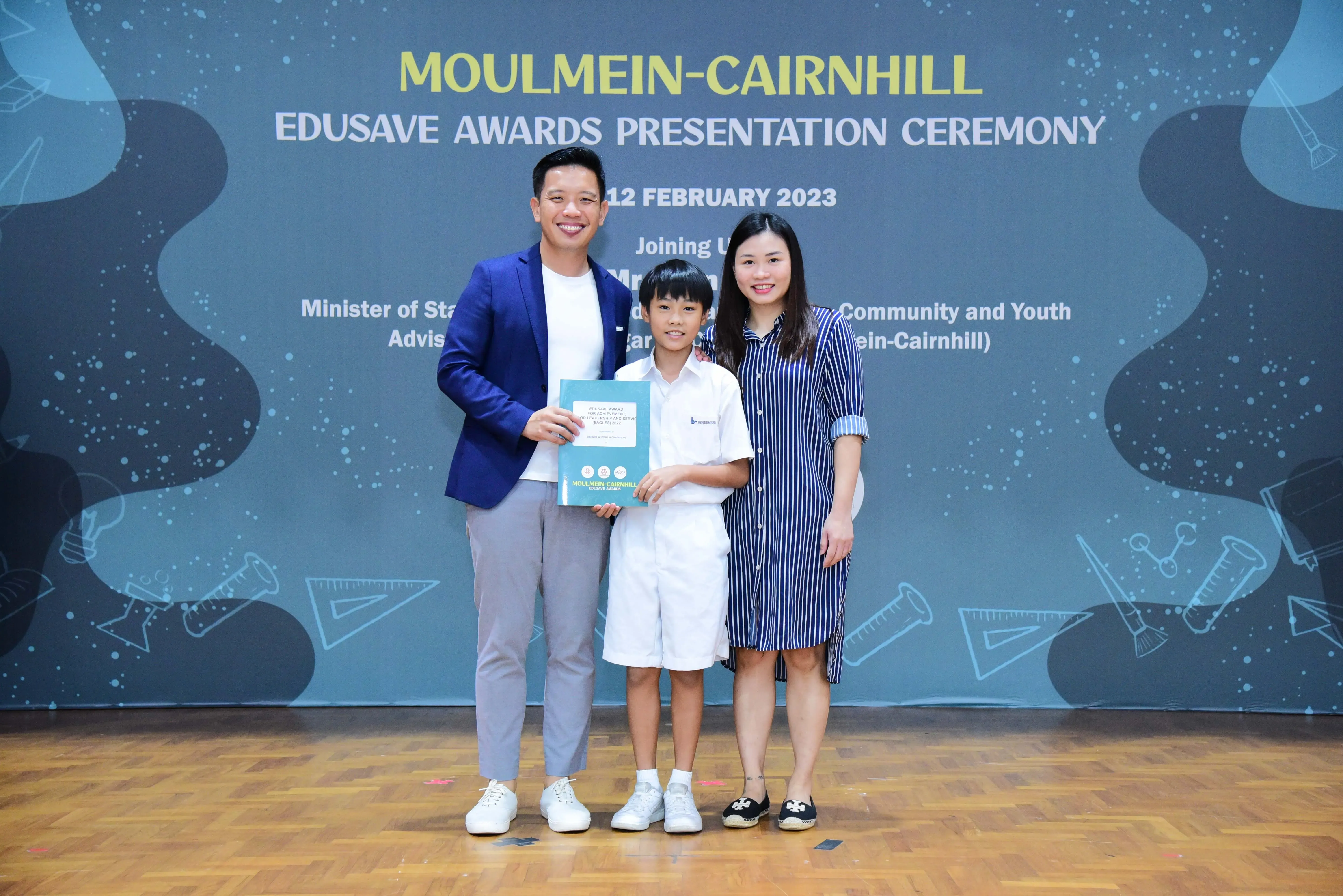 A boy in school uniform holds a certificate between two adults at the Moulmein-Cairnhill Edusave Awards Ceremony 2023.