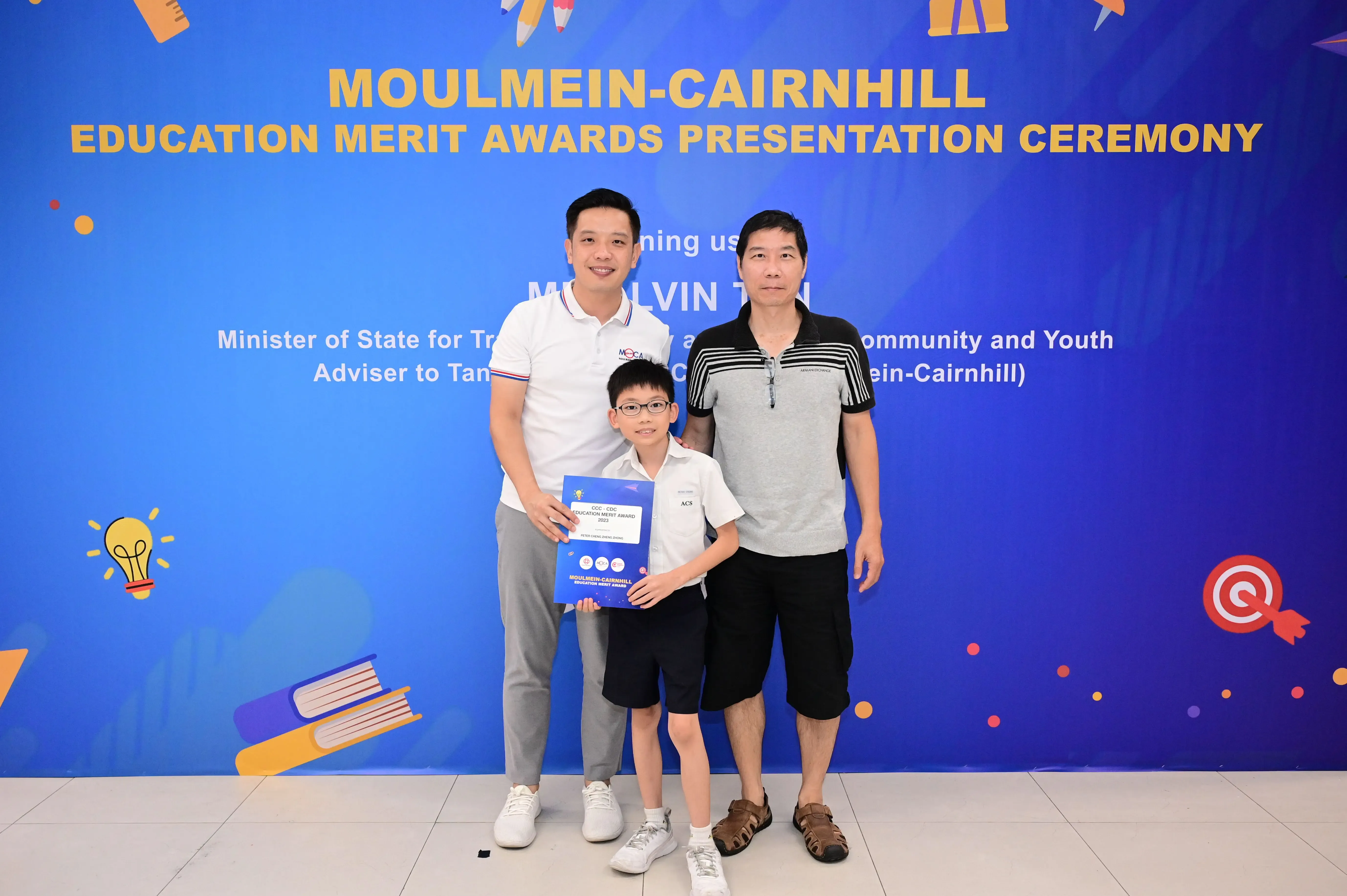 Two men and a young boy holding an award stand before a Moulmein-Cairnhill Education Merit Awards banner.